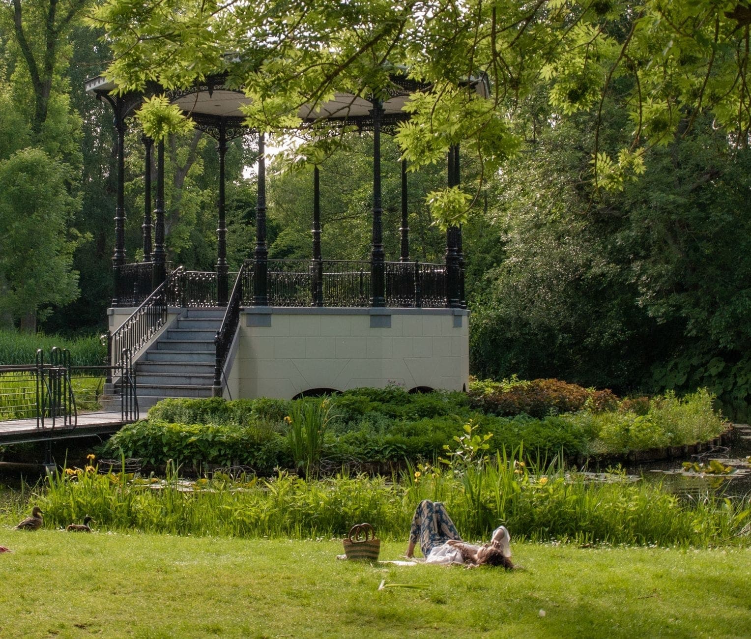 Person entspannt sich auf dem Gras in einem grünen Park mit einem dekorativen Pavillon und einem Teich, üppiges Grün schafft eine ruhige natürliche Umgebung.