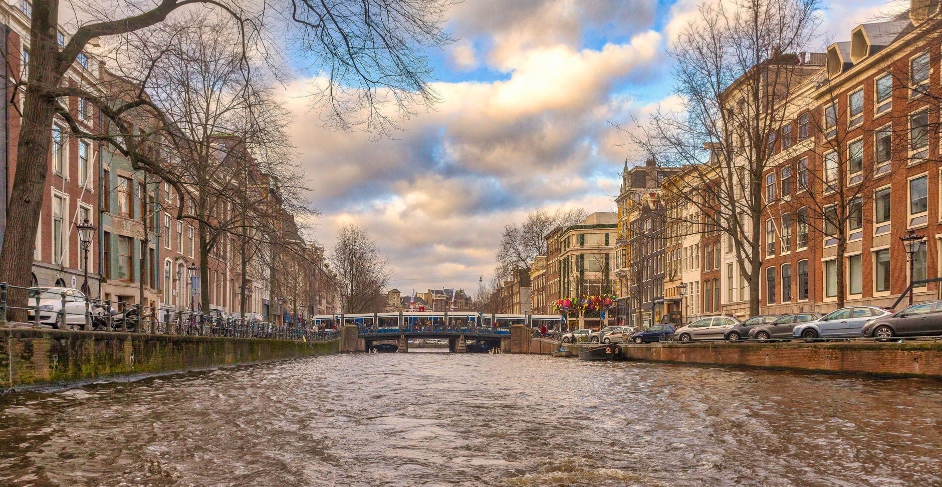 Weiter Blick auf einen Amsterdamer Kanal mit historischen Häusern, einer Brücke mit Straßenbahn und dramatischem Himmel mit Wolken.