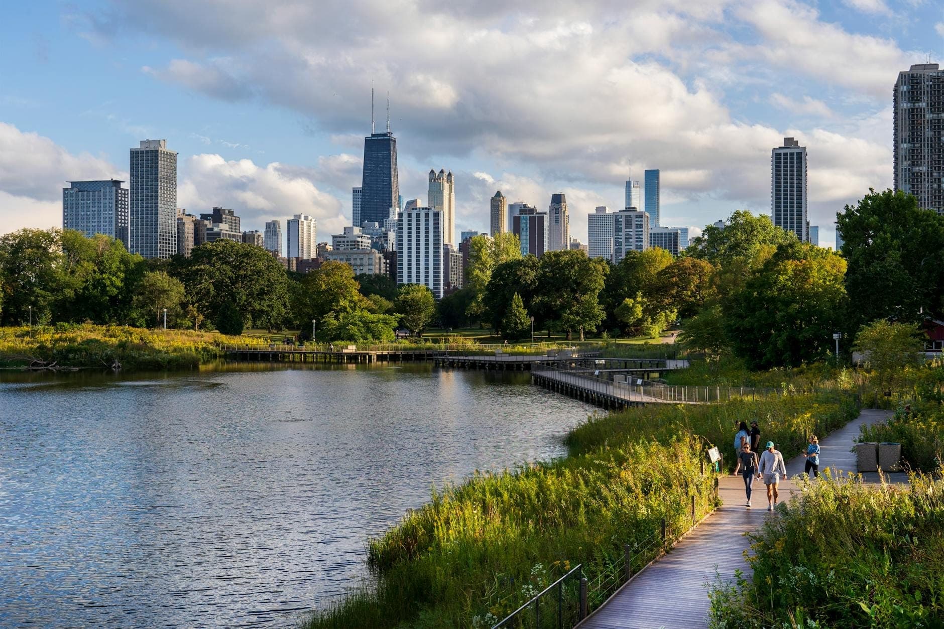 Chicago skyline viewed from a lakeside park with people walking on a boardwalk surrounded by greenery and water.