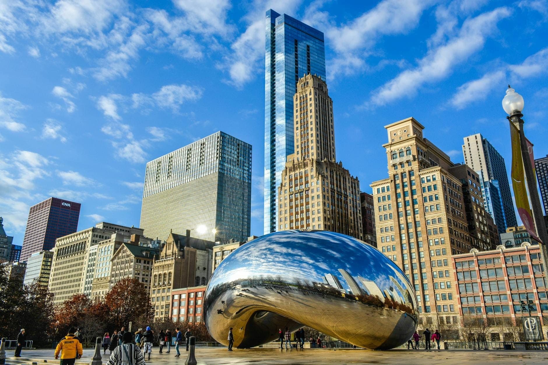 Cloud Gate sculpture, also known as The Bean, with people around it and the Chicago skyline in the background on a bright sunny day.