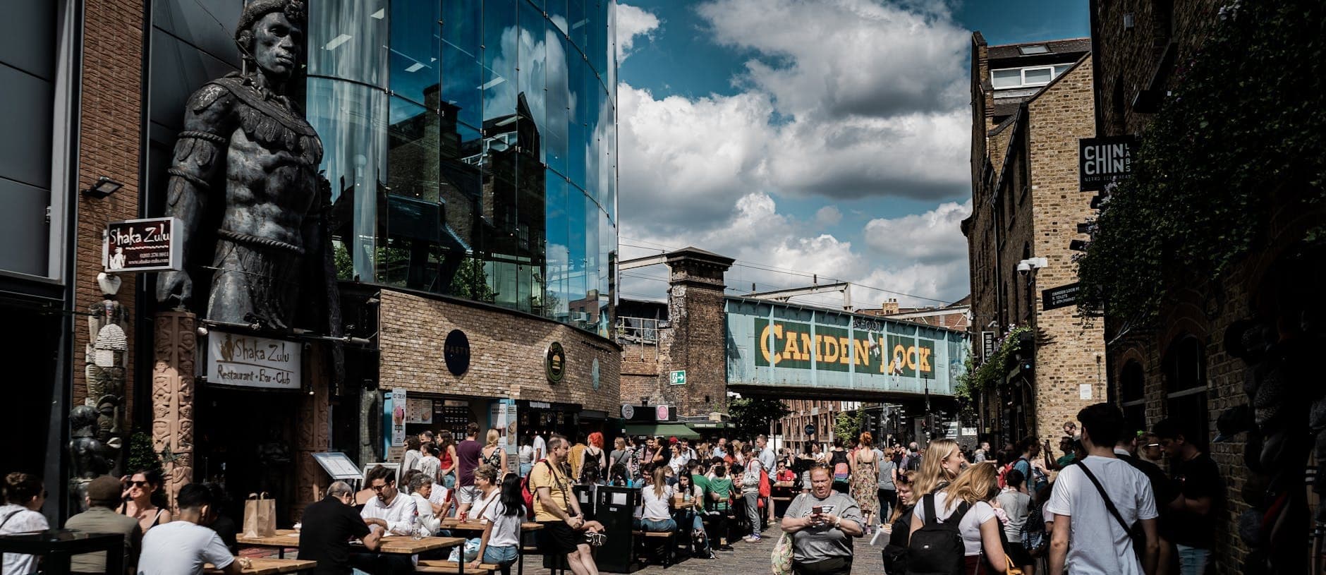 Geschäftiges Treiben auf dem Camden Market in London, mit Menschenmassen, Essensständen, Geschäften und der ikonischen Eisenbahnbrücke Camden Lock unter blauem Himmel.