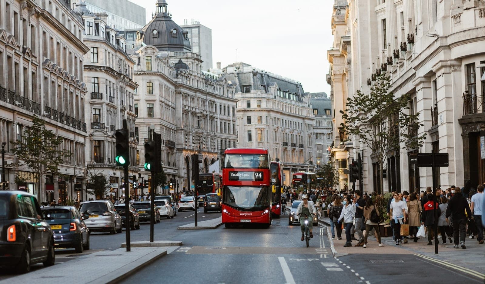 Belebte Londoner Straßenszene nahe dem Piccadilly Circus mit klassischen roten Doppeldeckerbussen, historischen Gebäuden, Autos und Fußgängern im sanften Nachmittagslicht.