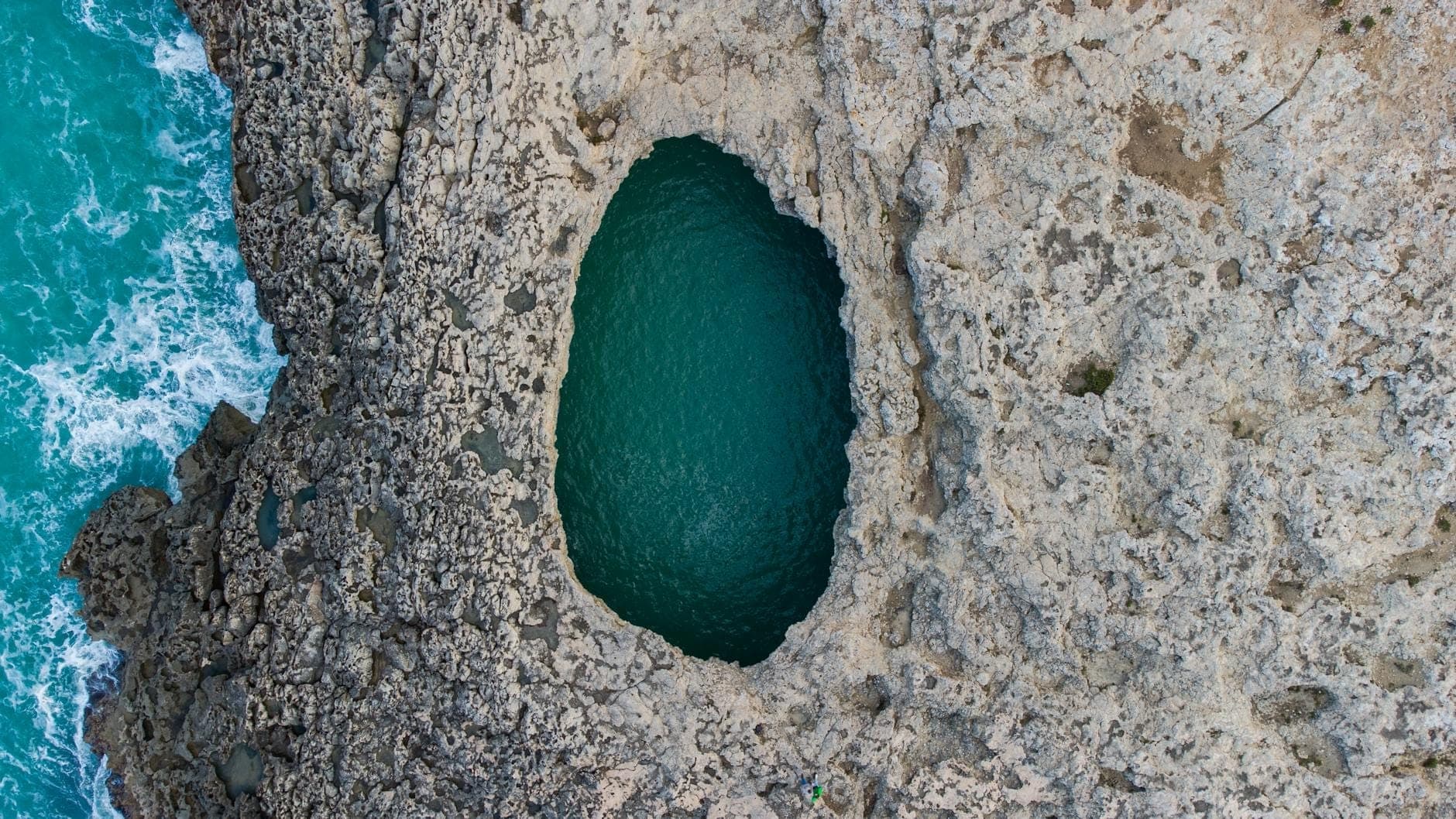 Luftaufnahme einer felsigen maltesischen Küste mit einem beeindruckenden natürlichen Schwimmloch neben dem türkisfarbenen Meer.