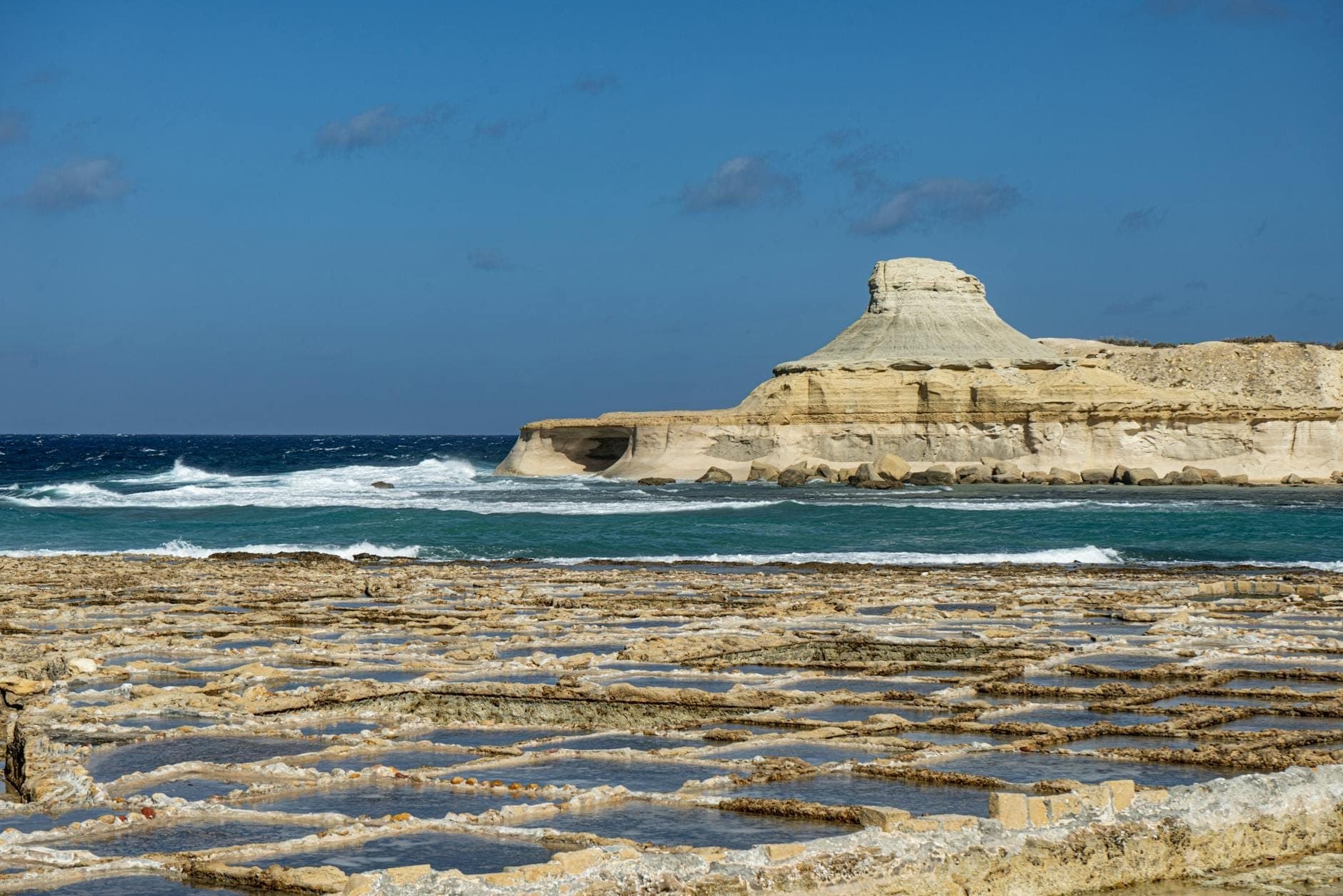 Küstenlinie bei Gozo mit ikonischen Salzgärten im Vordergrund und einem markanten Felshügel neben dem Mittelmeer unter einem klaren blauen Himmel.