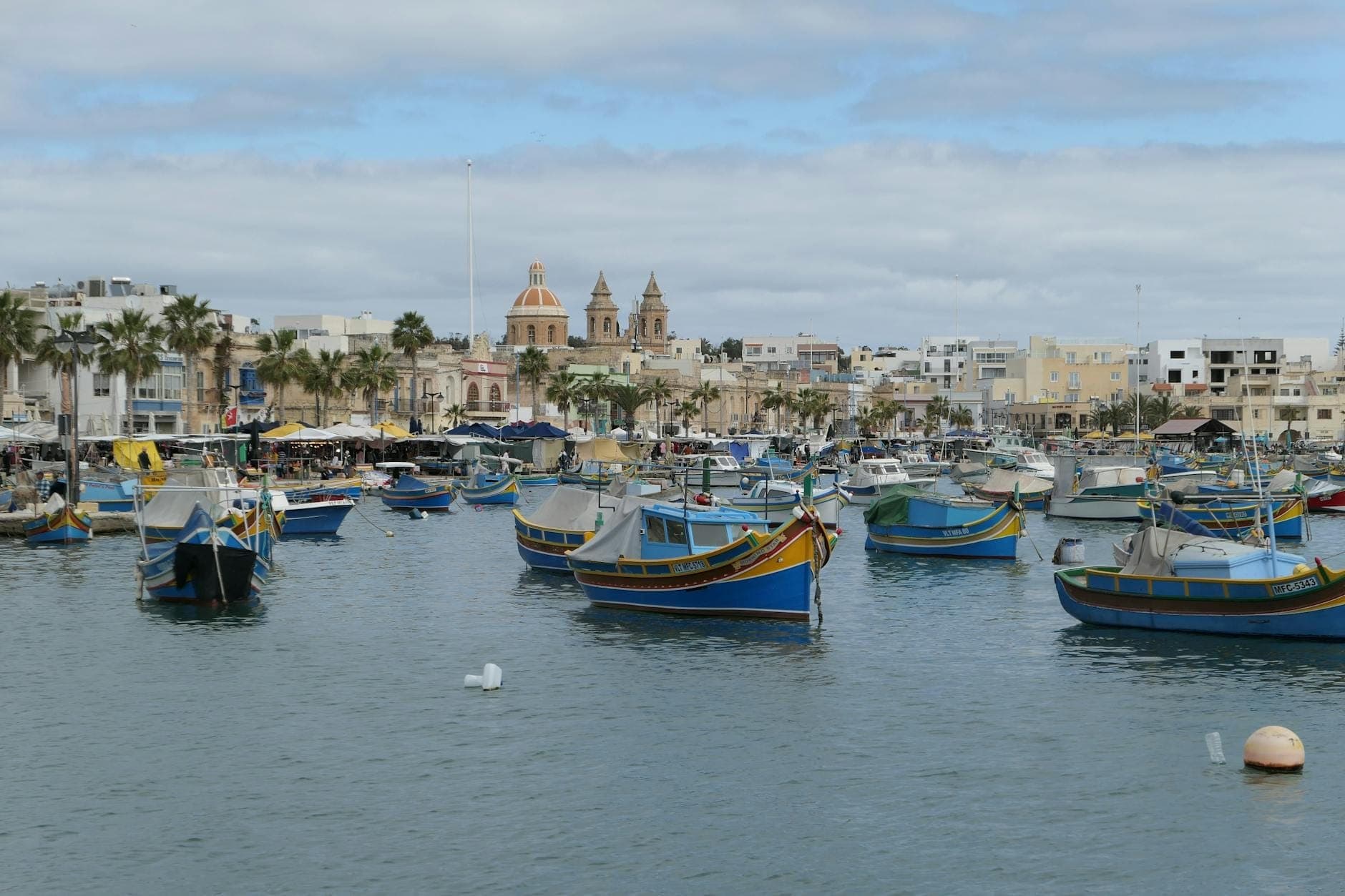 Bunte Fischerboote in einem maltesischen Hafen mit einer traditionellen Kirche und Marktständen im Hintergrund unter einem bewölkten Himmel.