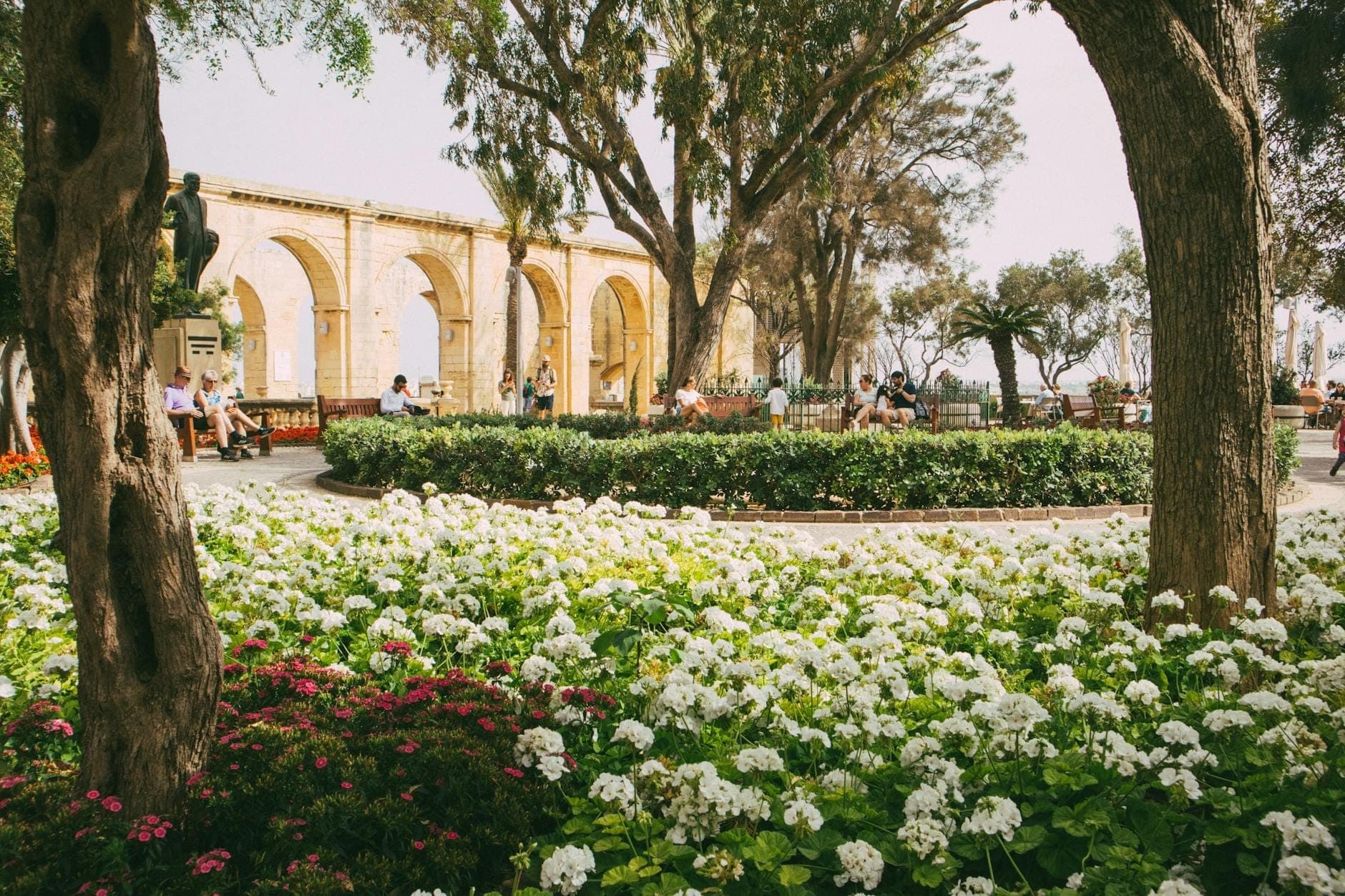 Blumengefüllter öffentlicher Garten mit großen Steinbögen, Bäumen und Menschen, die auf Bänken entspannen, in Malta.