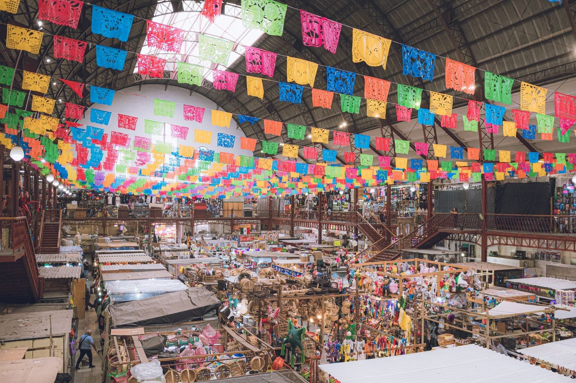 Brightly lit indoor Mexican market scene with colorful papel picado banners overhead and bustling vendor stalls below.