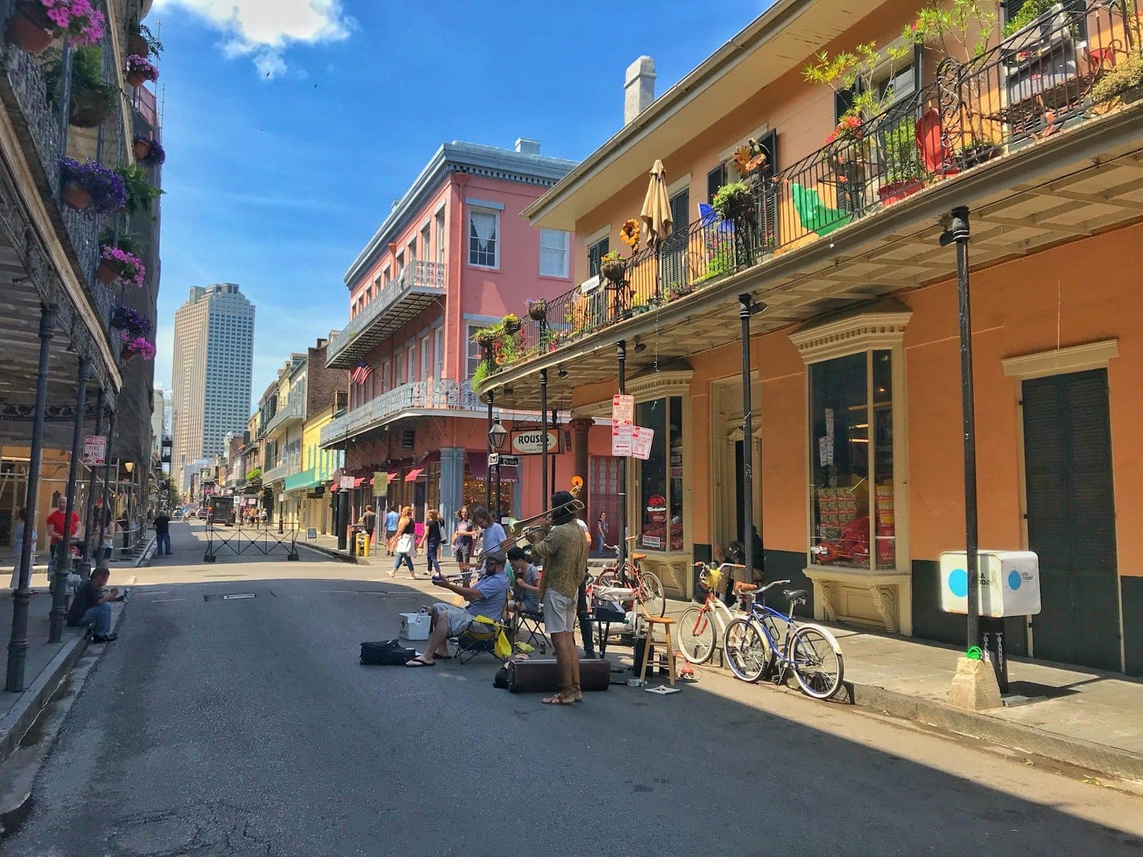 Des bâtiments historiques colorés et des balcons en fer forgé longent une rue animée de New Orleans, avec des musiciens jouant pour les passants par une journée ensoleillée.