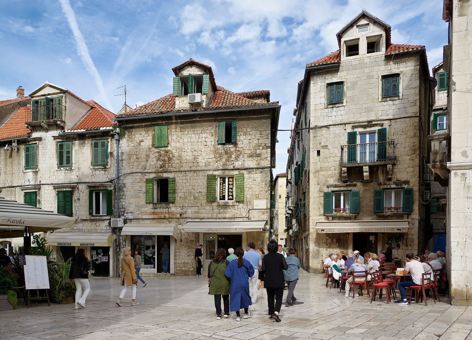 La gente passeggia in una piazza soleggiata nel centro storico di Split, circondata da edifici in pietra con persiane verdi e tetti in terracotta.