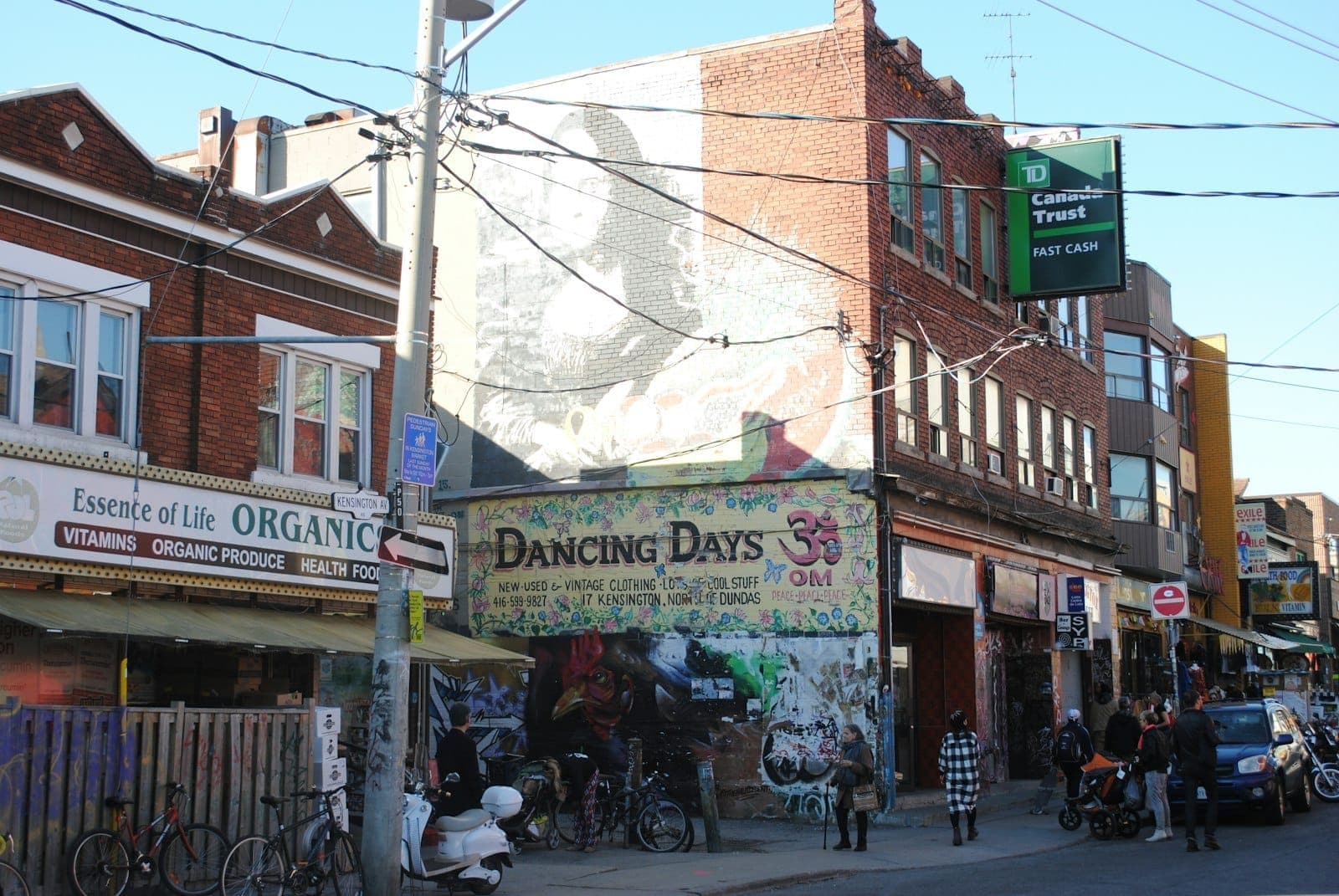 Vista della strada del Kensington Market a Toronto, con vivaci negozi indipendenti, pedoni e un colorato murales in una giornata di sole.