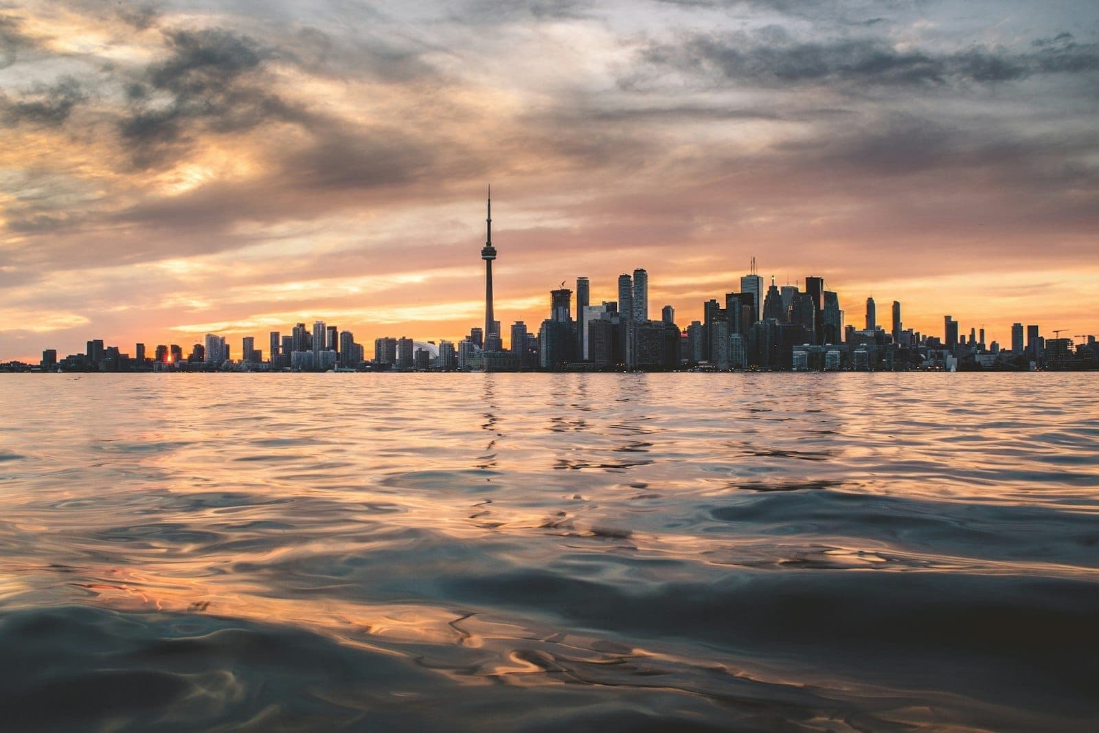 Cakrawala Toronto saat matahari terbenam dengan awan dramatis, CN Tower terlihat jelas, gedung-gedung kota terpantul di permukaan danau yang tenang di latar depan.