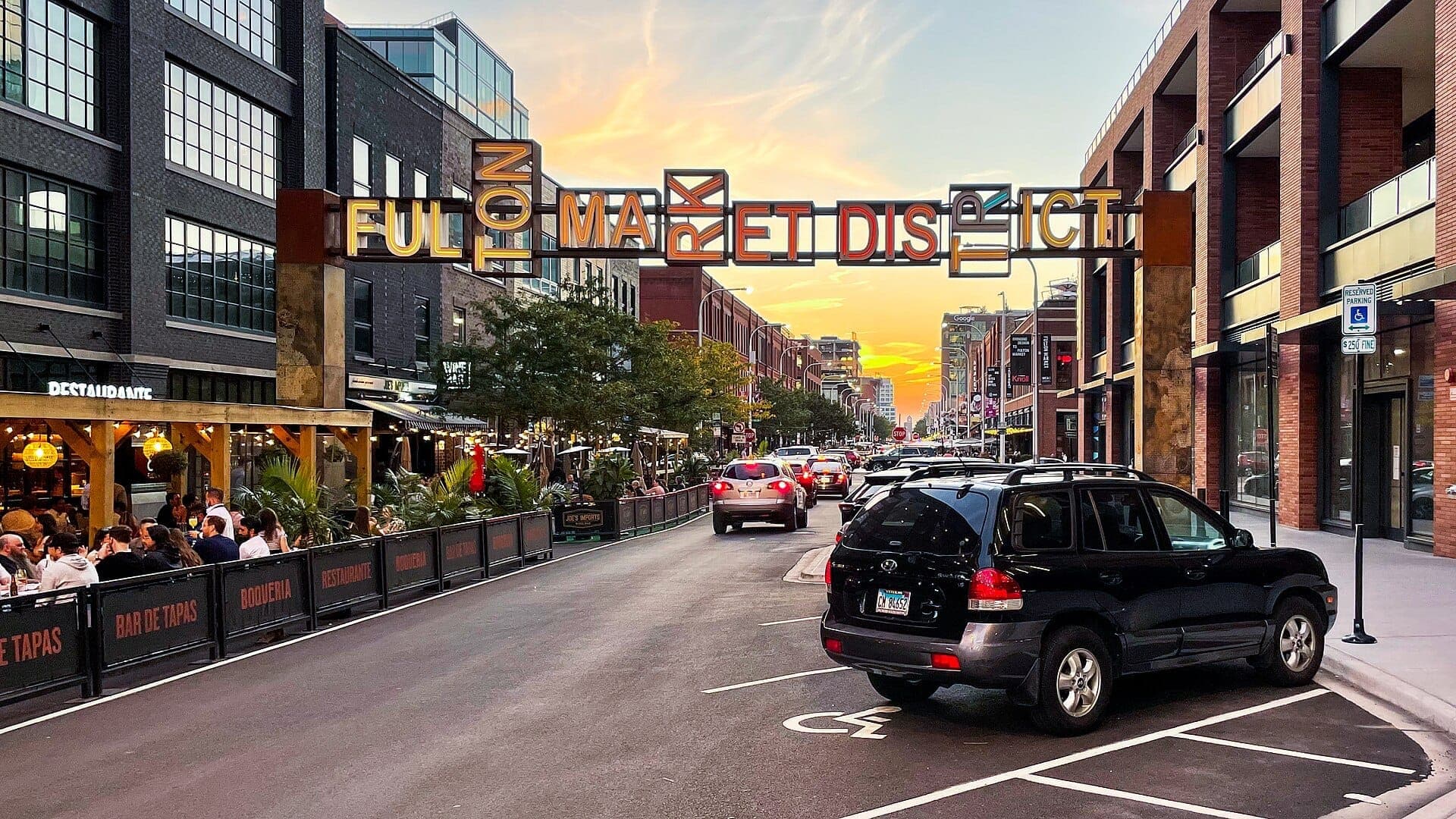 ป้ายทางเข้า Fulton Market District พร้อมถนนคึกคัก อาคารอิฐ พื้นที่รับประทานอาหารกลางแจ้ง รถยนต์ และท้องฟ้ายามพระอาทิตย์ตกที่สดใสใน Chicago