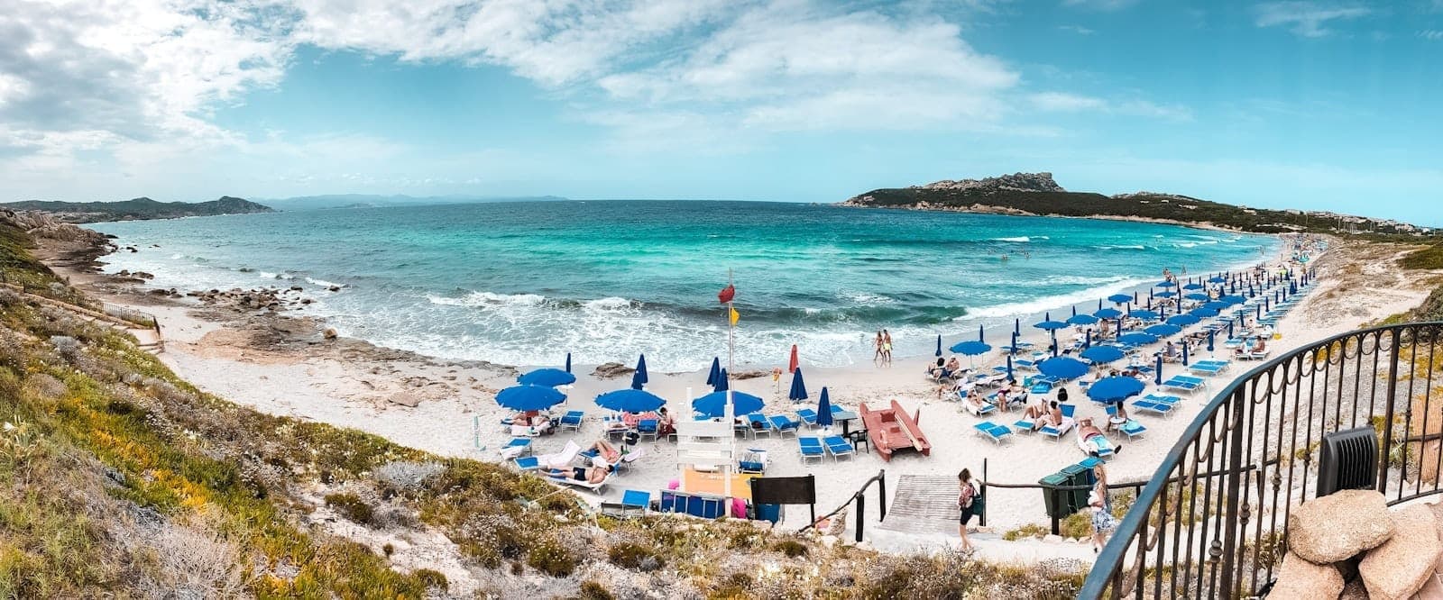 Vista panorámica de la playa de Gallura en Cerdeña con agua turquesa, arena blanca, costa rocosa y filas de sombrillas y tumbonas azules.