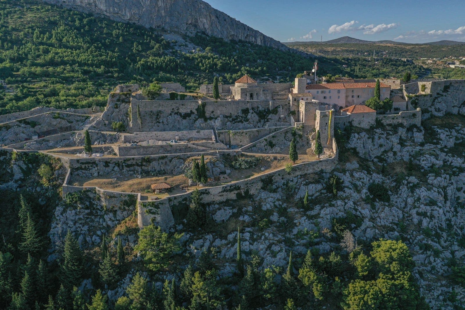 Vista aerea della Fortezza di Klis arroccata in cima a una collina rocciosa, circondata da mura in pietra ed edifici storici, con colline verdi sullo sfondo.