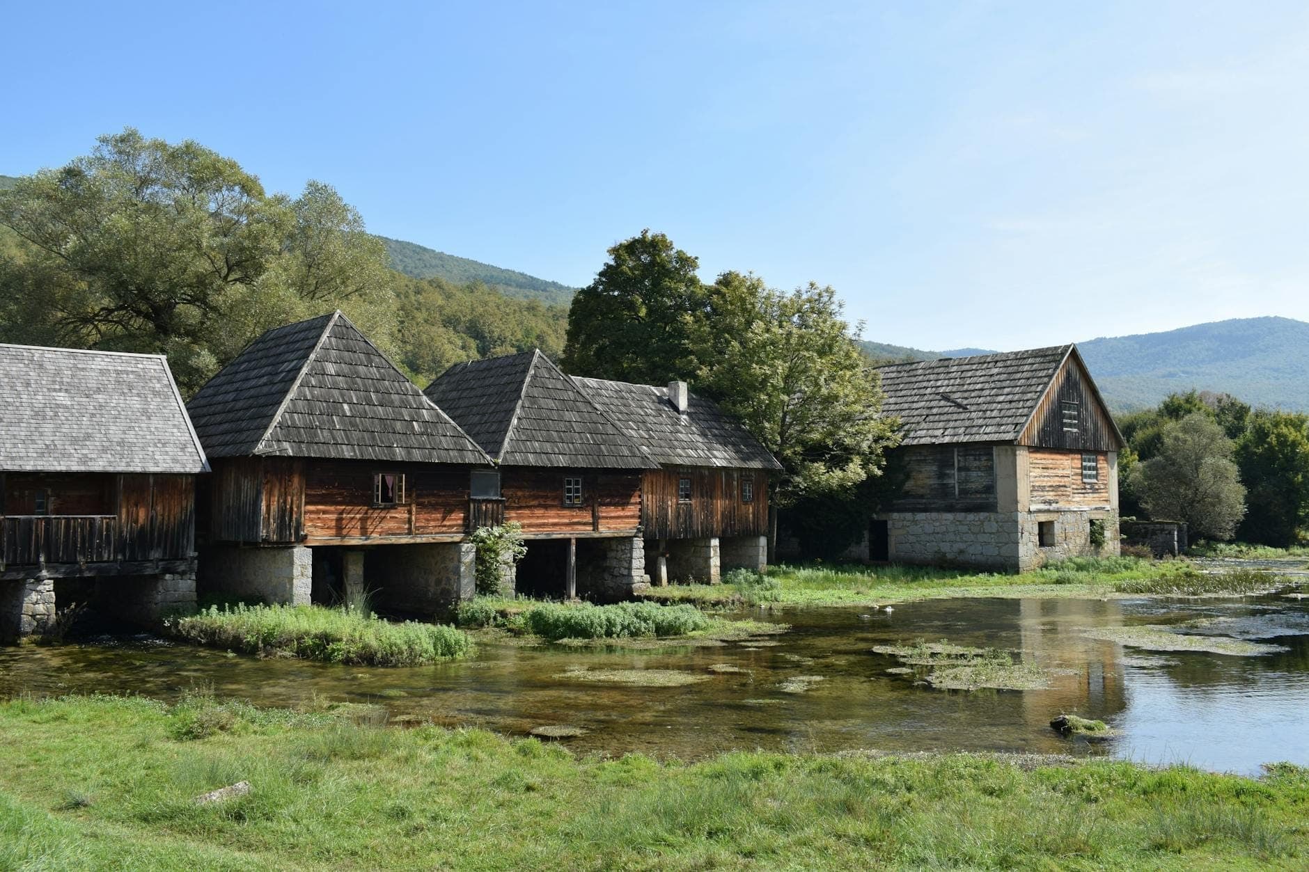 Fila di vecchi mulini fluviali in legno con basi in pietra lungo una riva erbosa sotto un cielo sereno vicino a Spalato, Croazia.