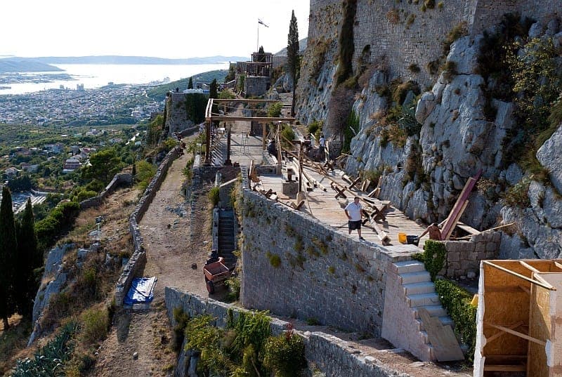 Vista panoramica della Fortezza di Klis in Croazia con mura in pietra, scogliere frastagliate, costruzioni del set cinematografico e la città di Split visibile in lontananza.