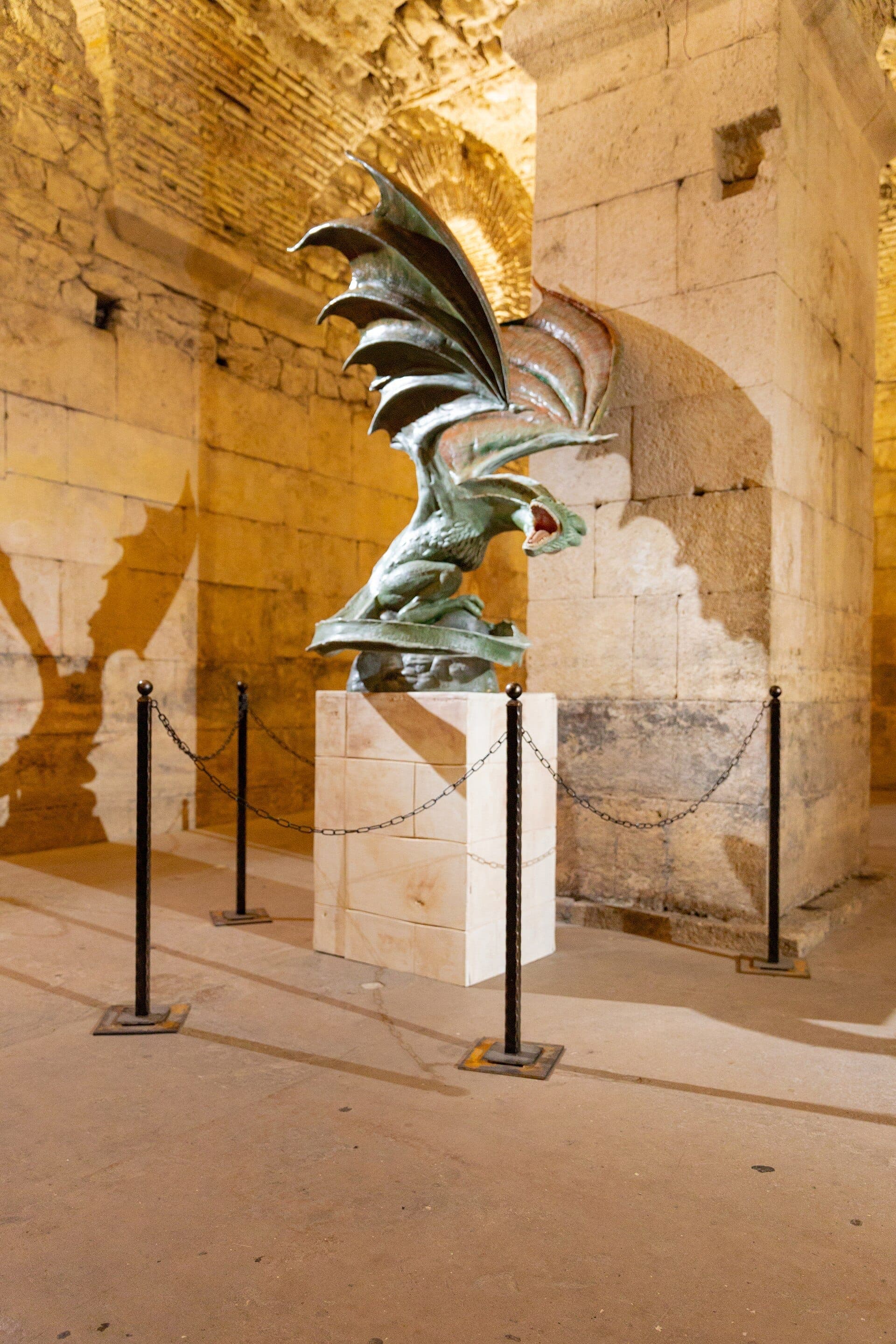 Una scultura di drago esposta su un piedistallo all'interno del Game of Thrones Museum Split con pareti in pietra, circondata da catene e un'illuminazione suggestiva.