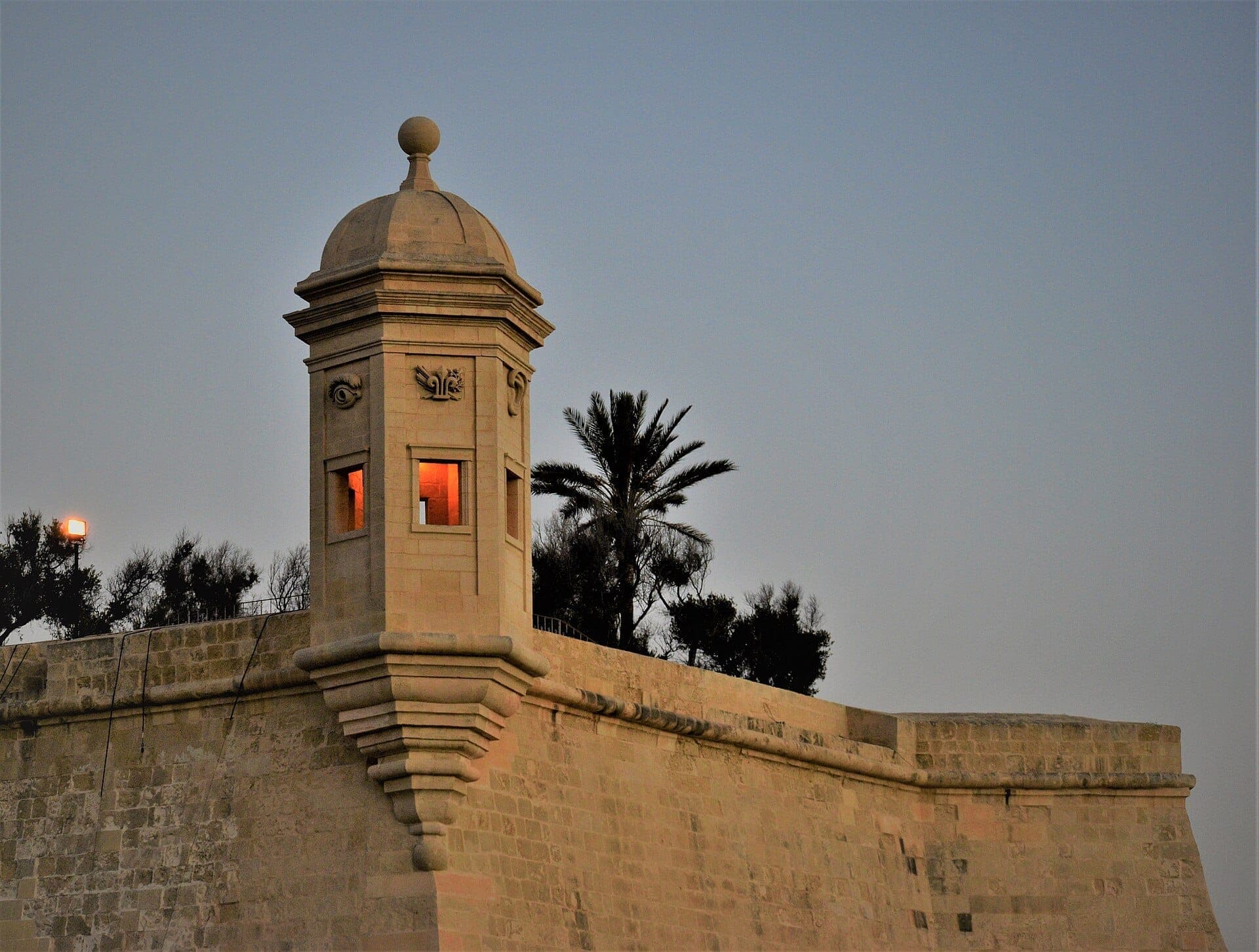 Steinerner Wachturm in den Gardjola Gardens in Senglea, Malta, mit warmem Abendlicht und Palmen als Silhouette vor klarem Himmel.