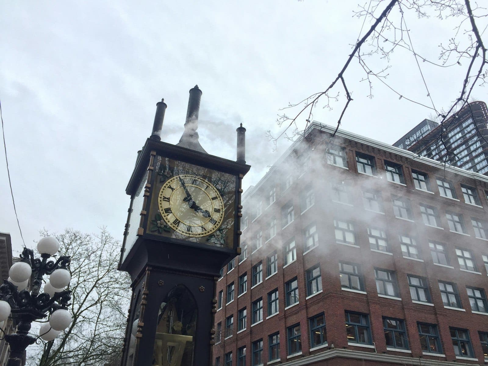 นาฬิกาไอน้ำ Gastown Steam Clock พ่นไอน้ำในวันที่มีเมฆมาก ล้อมรอบด้วยอาคารอิฐประวัติศาสตร์และกิ่งไม้ไร้ใบในย่านใจกลางเมือง Vancouver
