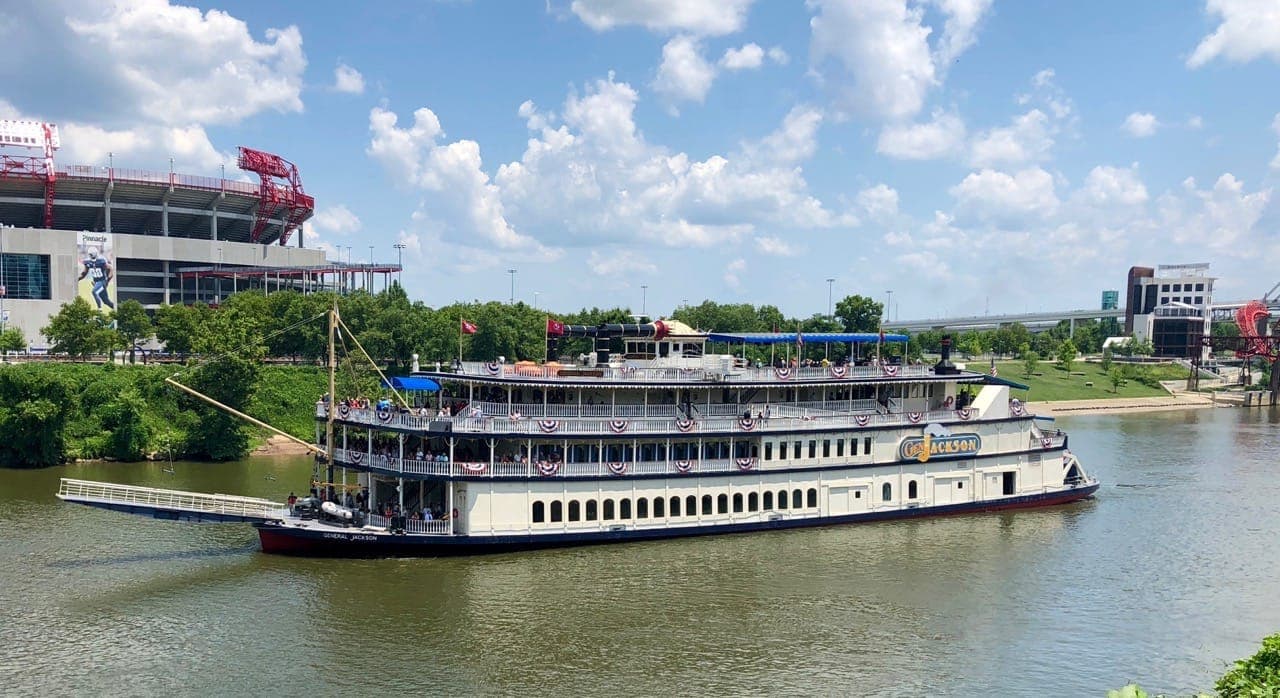 The General Jackson Showboat, decorated with American bunting, cruises along the Cumberland River under a partly cloudy sky with a stadium in the background.