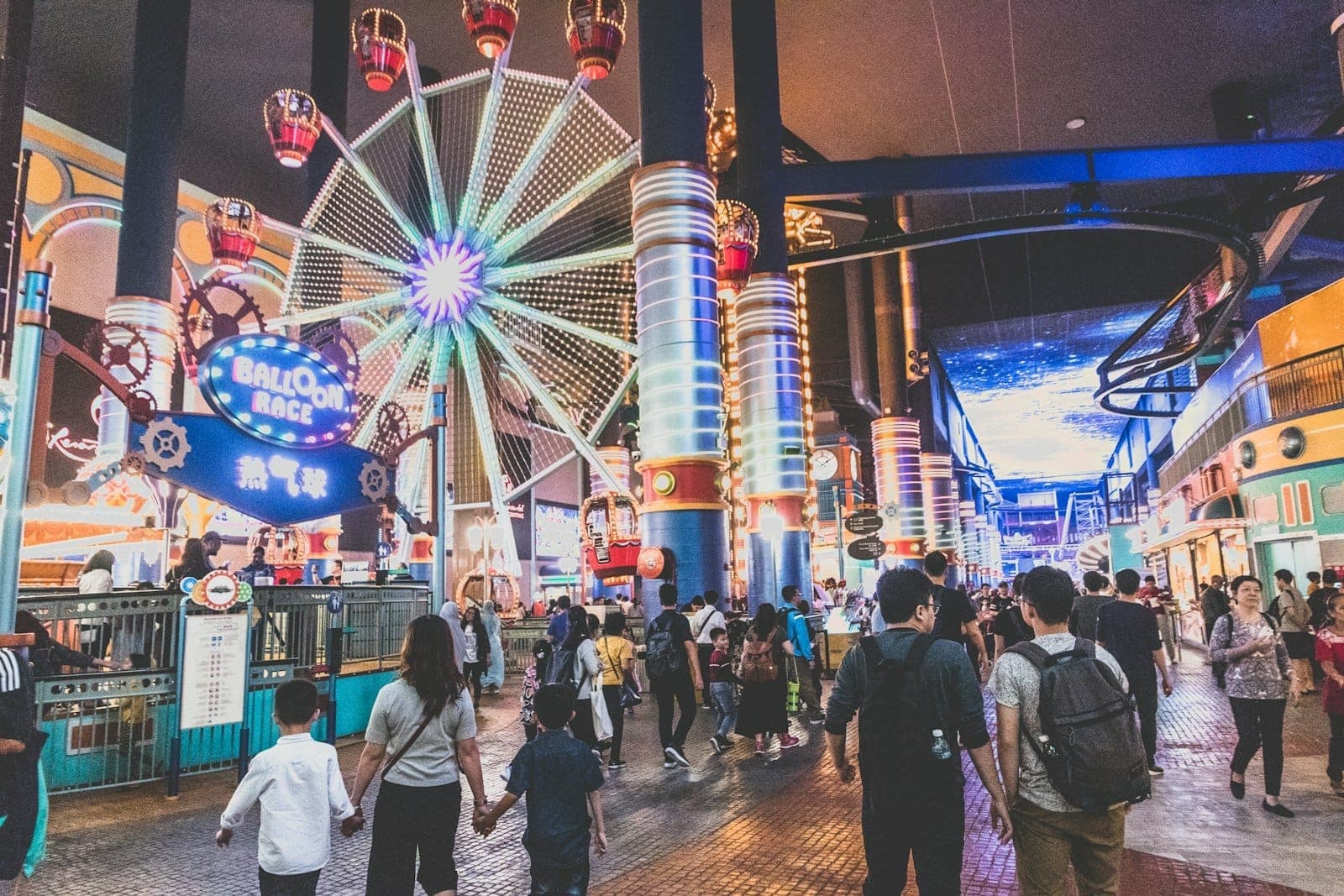 Indoor amusement park at Genting Highlands with a brightly lit Ferris wheel, Balloon Race ride, and families walking under colorful lights.