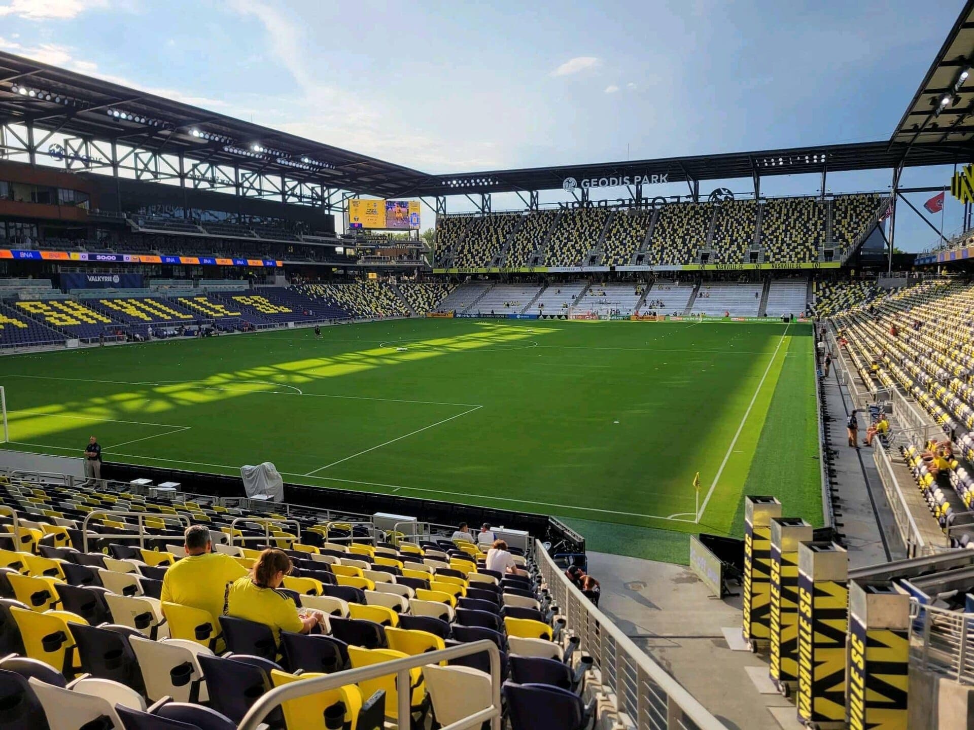 Wide view of GEODIS Park stadium in Nashville, showing yellow and black seats, green soccer field, and a few spectators in sunlight.