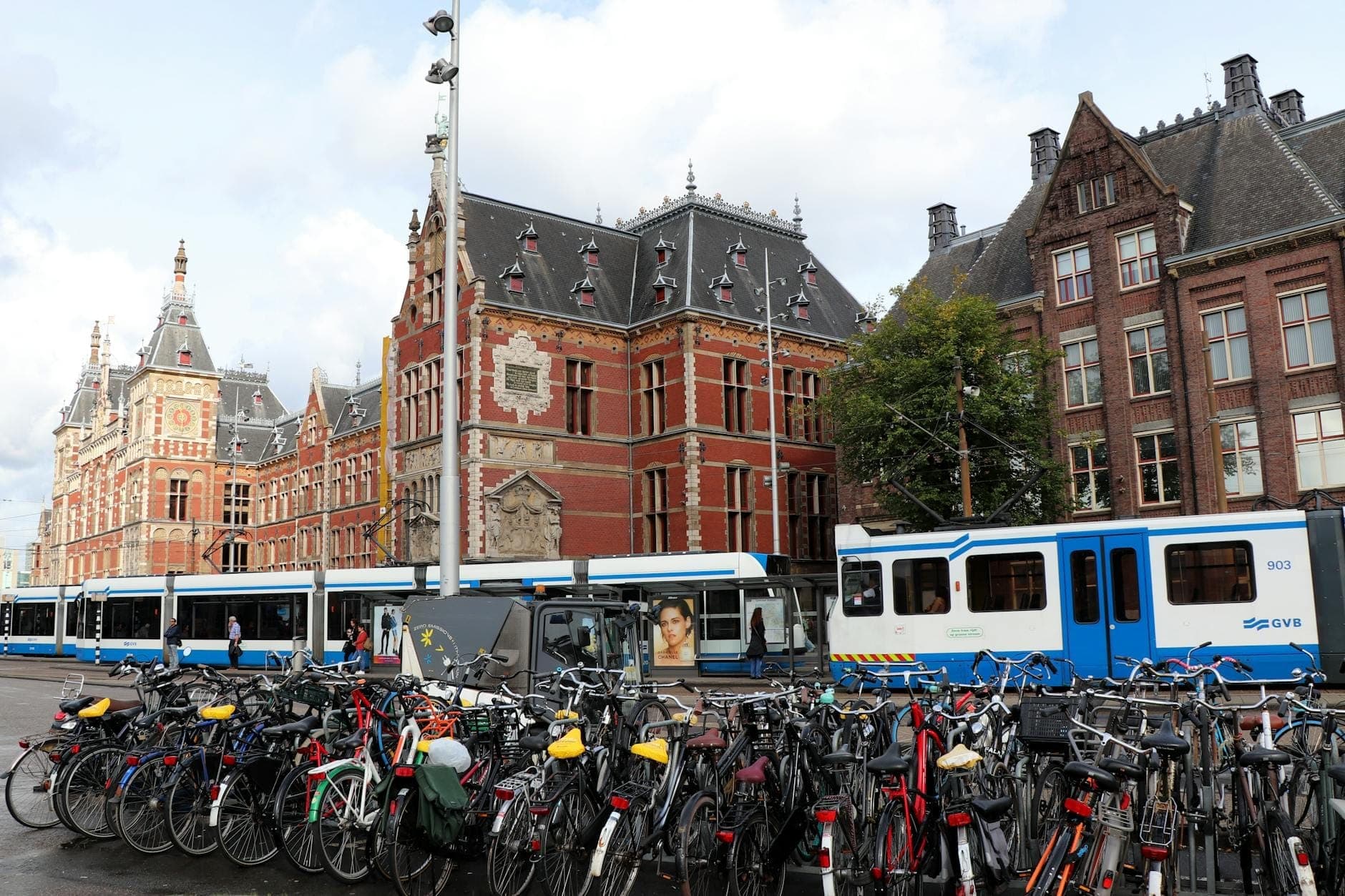 Bahnhof Amsterdam Centraal mit davor geparkten Fahrrädern und vorbeifahrenden blau-weißen Straßenbahnen, die die wichtigsten städtischen Verkehrsmittel gemeinsam veranschaulichen.