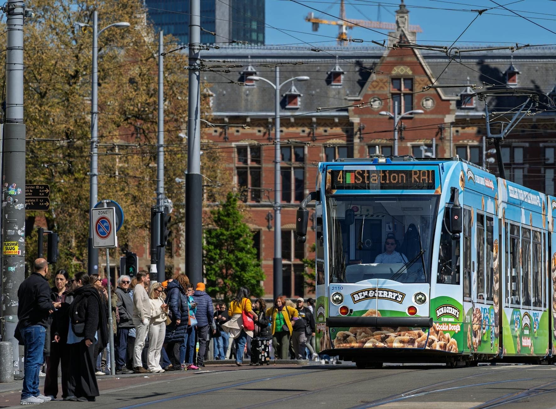 Amsterdamer Straßenbahn an einem belebten Bahnsteig mit einer großen Menschenmenge, die vor der traditionellen Stadtarchitektur an einem sonnigen Tag wartet.