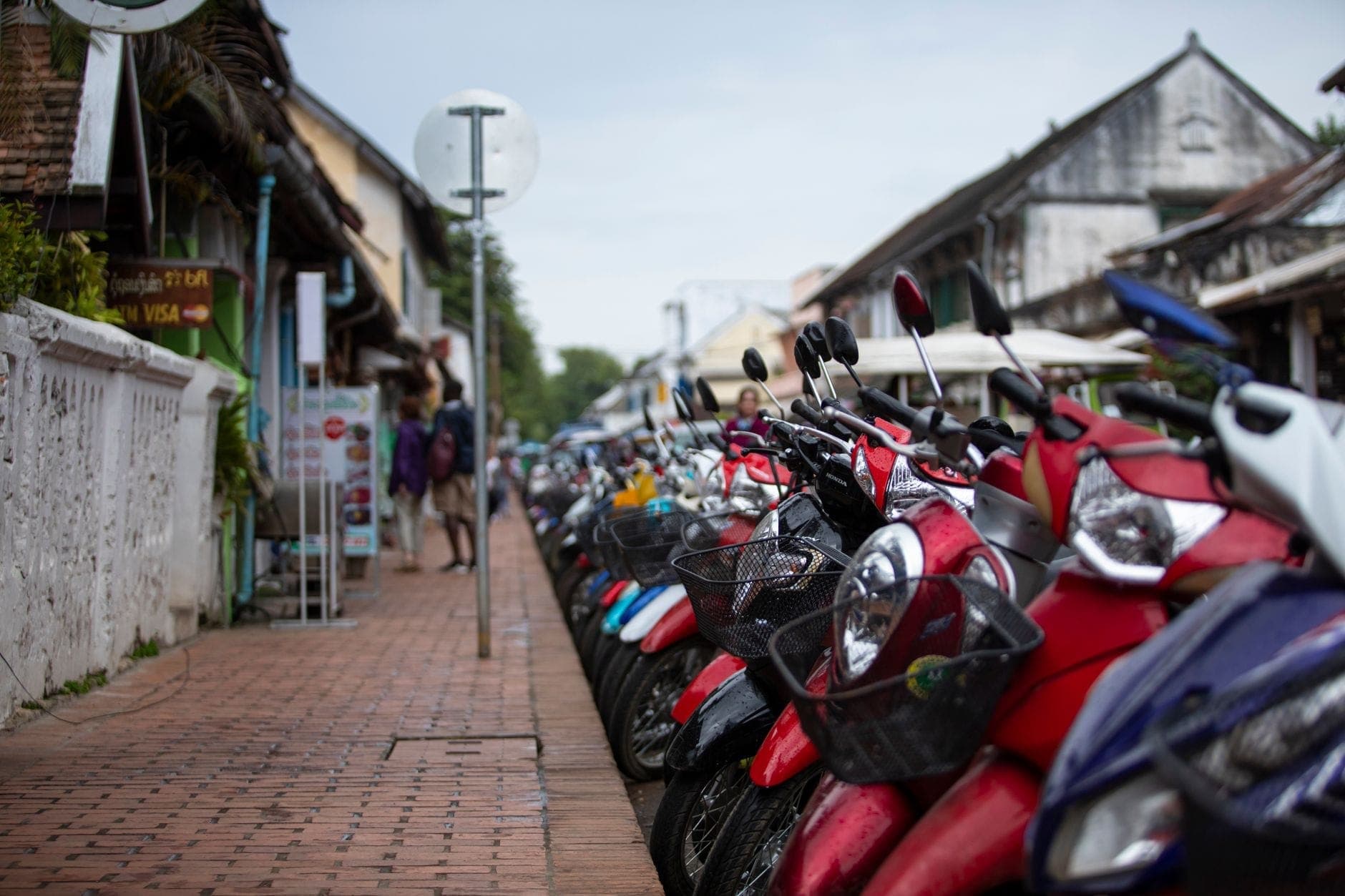 Güneydoğu Asya'daki bir şehirde kaldırım boyunca park edilmiş scooter sırası, binalar ve sokak tabelaları görünüyor.