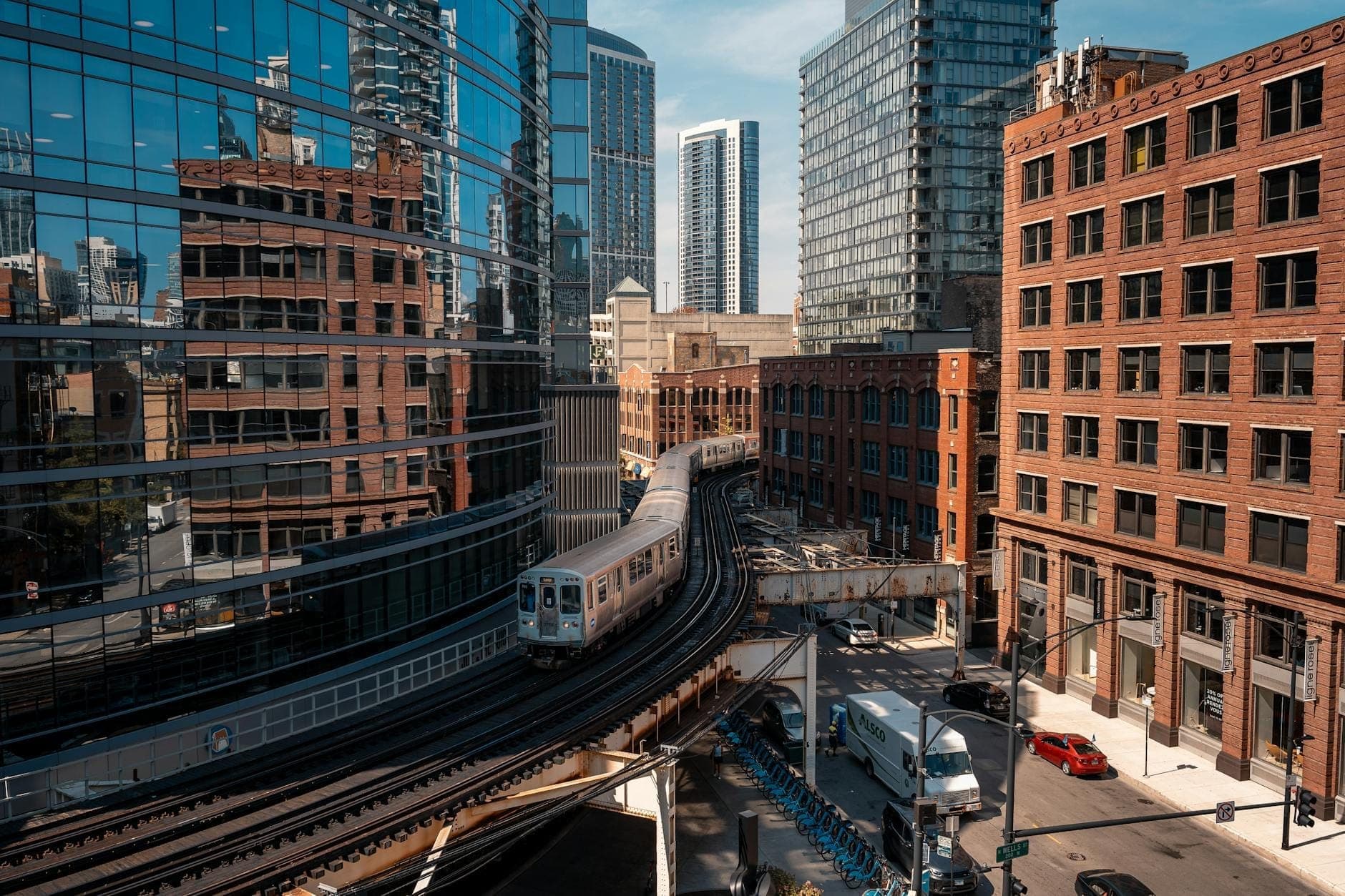 A Chicago 'L' train curves through downtown skyscrapers on elevated tracks, showcasing urban architecture, busy streets, and iconic public transit on a clear day.
