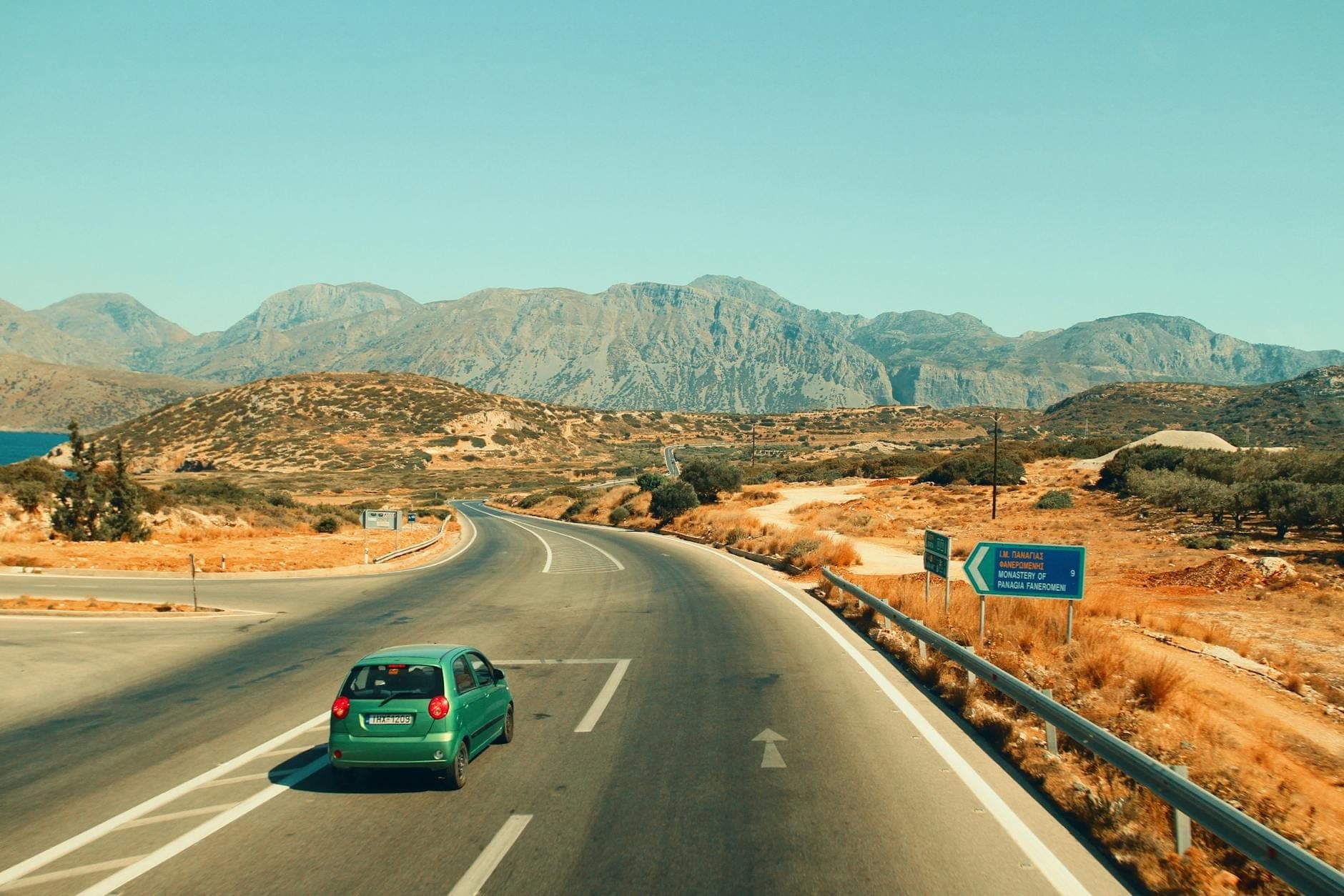 Auto fährt auf einer Autobahn durch eine trockene, bergige Landschaft mit griechischen Straßenschildern und den Bergen Kretas im Hintergrund