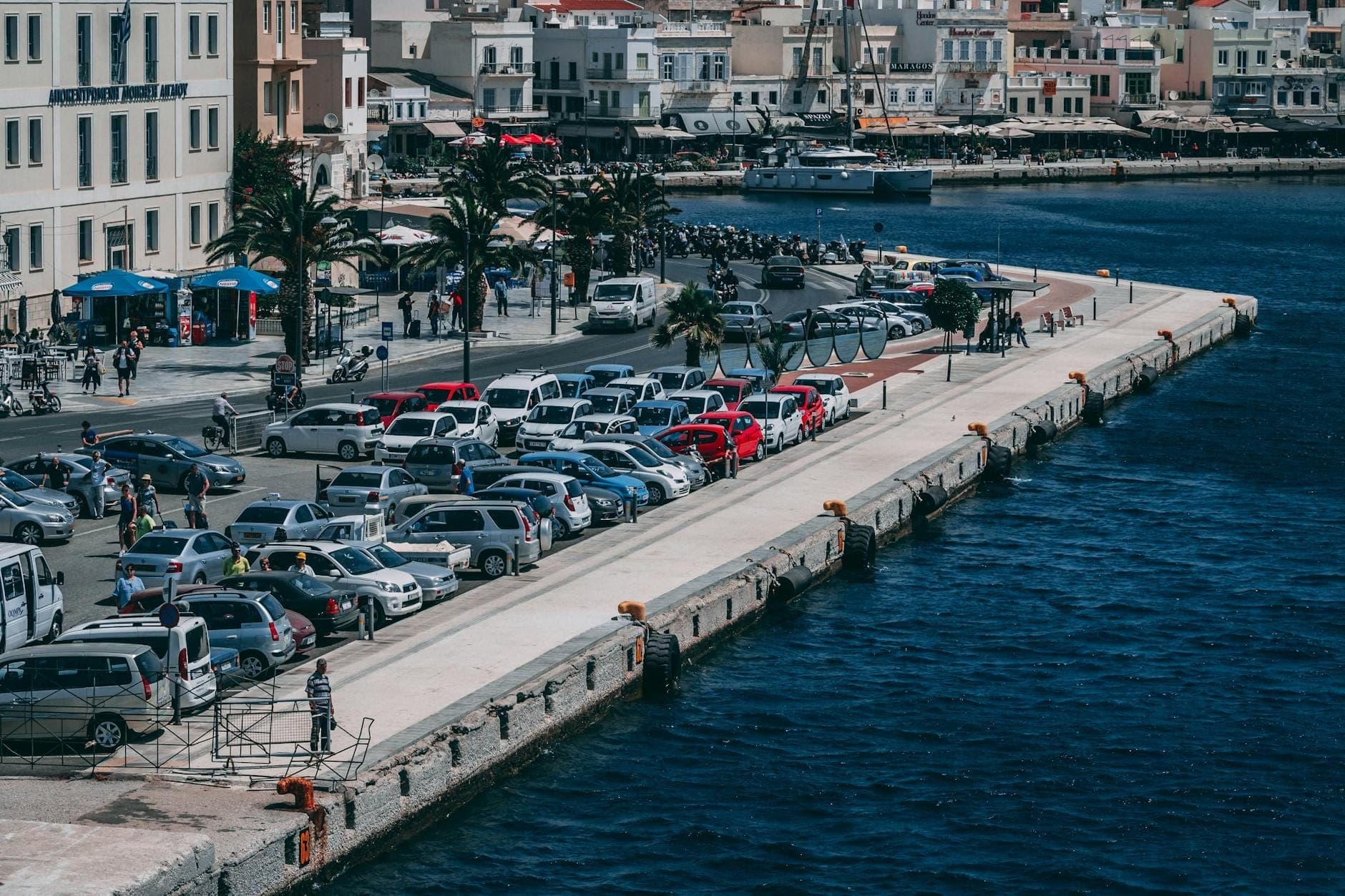 Ein großer Parkplatz voller Autos neben einem malerischen Hafen auf Kreta, mit Gebäuden und Palmen entlang der Uferpromenade.