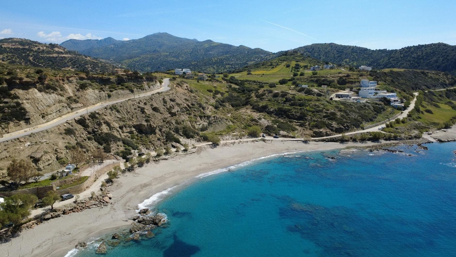 Eine kurvenreiche Küstenstraße entlang zerklüfteter Hügel und einem Strand, mit vereinzelten Gebäuden und Bergen in der Ferne auf Kreta.