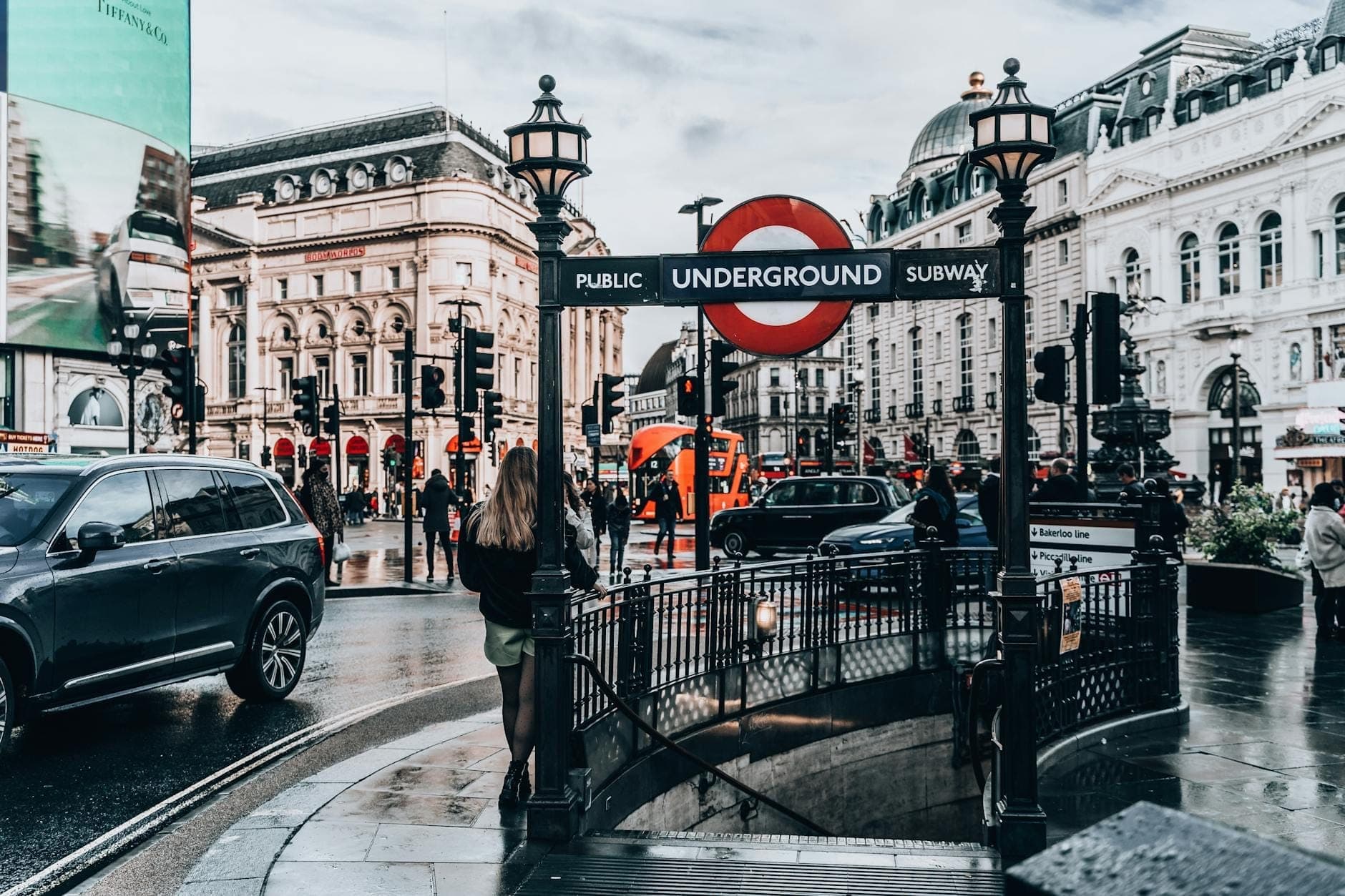 Вход на станцию London Underground на уровне улицы на Piccadilly Circus, с культовым знаком roundel и оживлённой городской сценой при дневном свете.