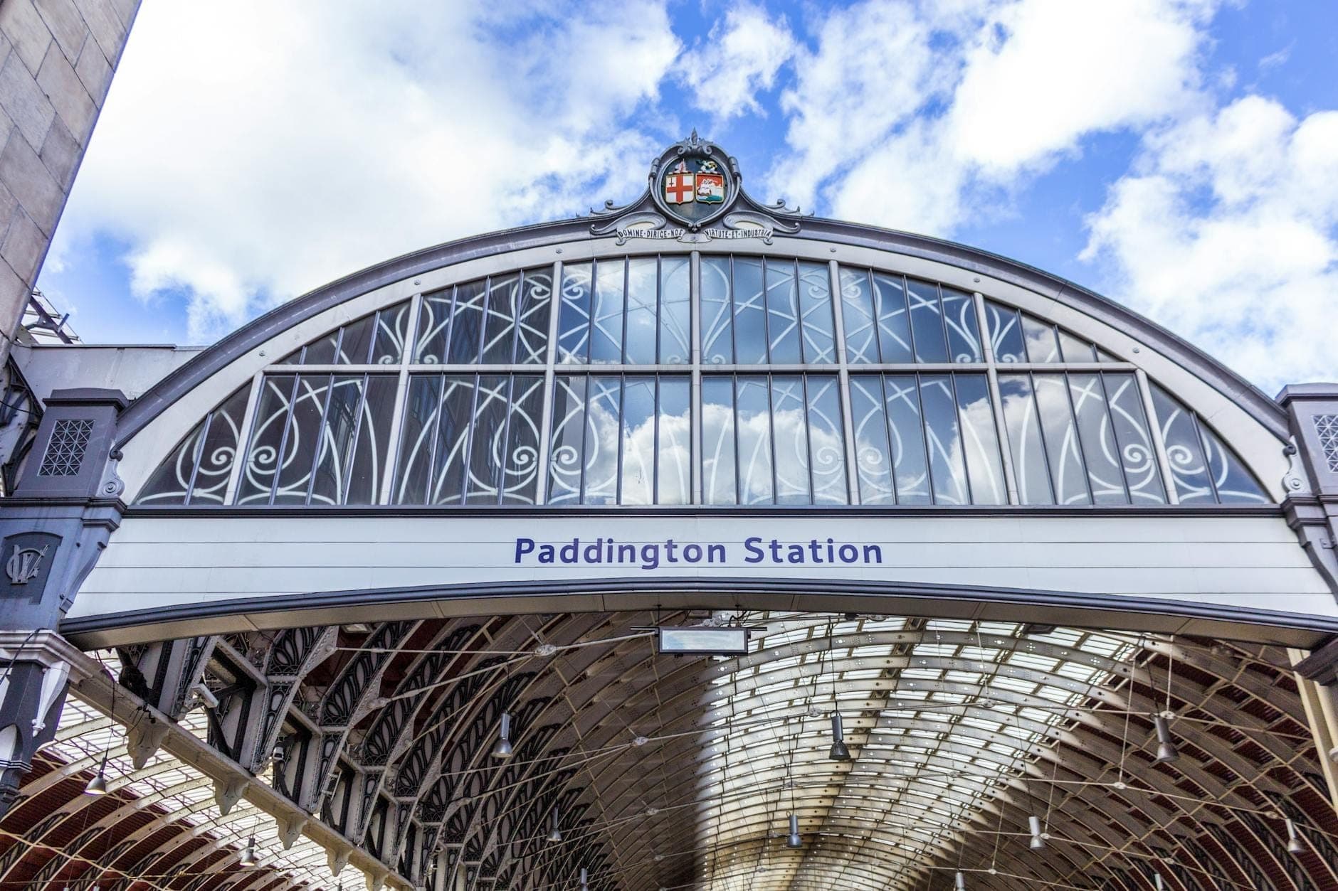 Внешний вход на London Paddington Station с вывеской и арочной стеклянной крышей в ясный день с голубым небом и облаками.