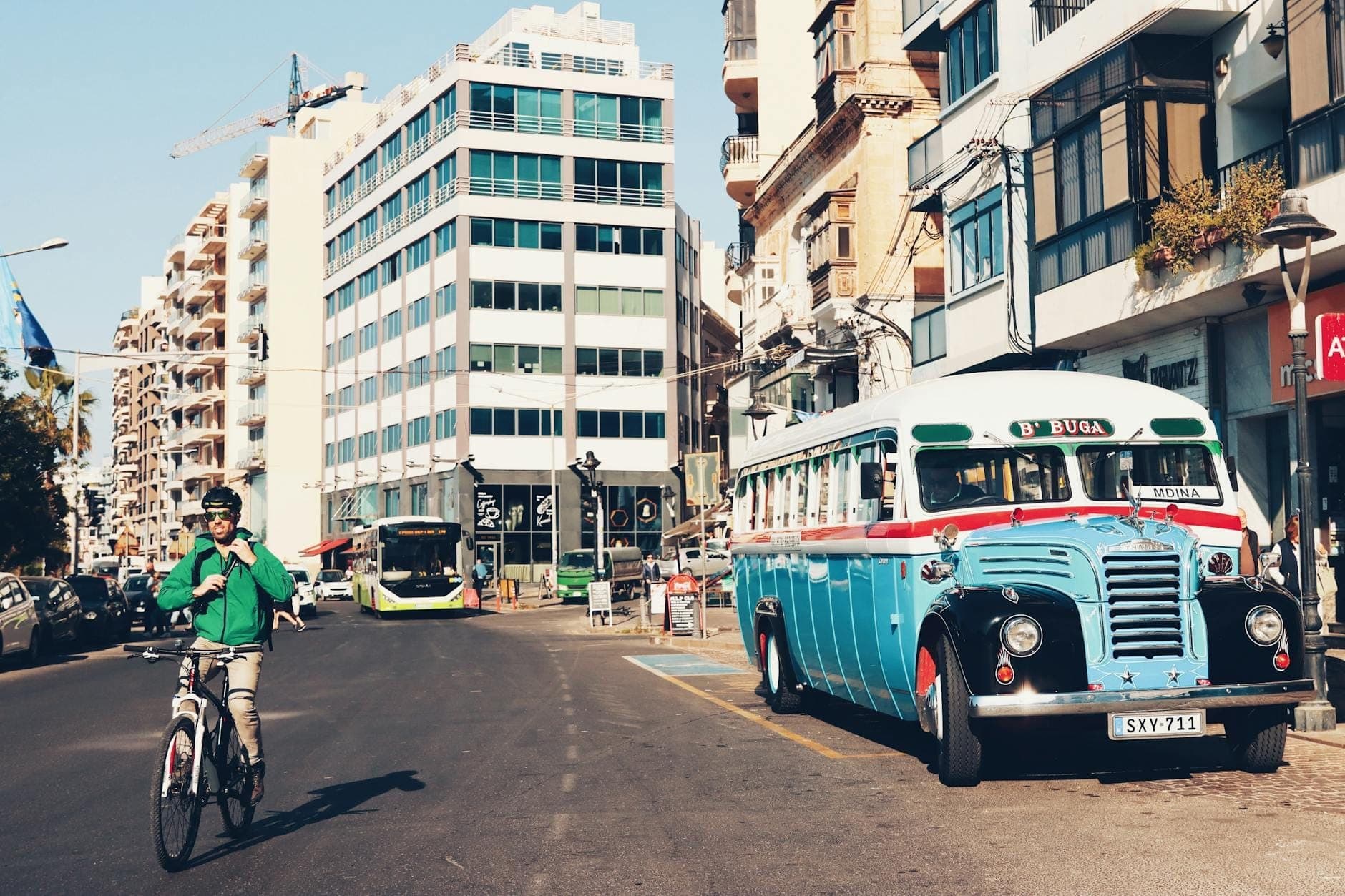 Autobús maltés clásico estacionado en una calle de la ciudad, con edificios modernos y un ciclista en primer plano, mostrando el entorno urbano de Malta.