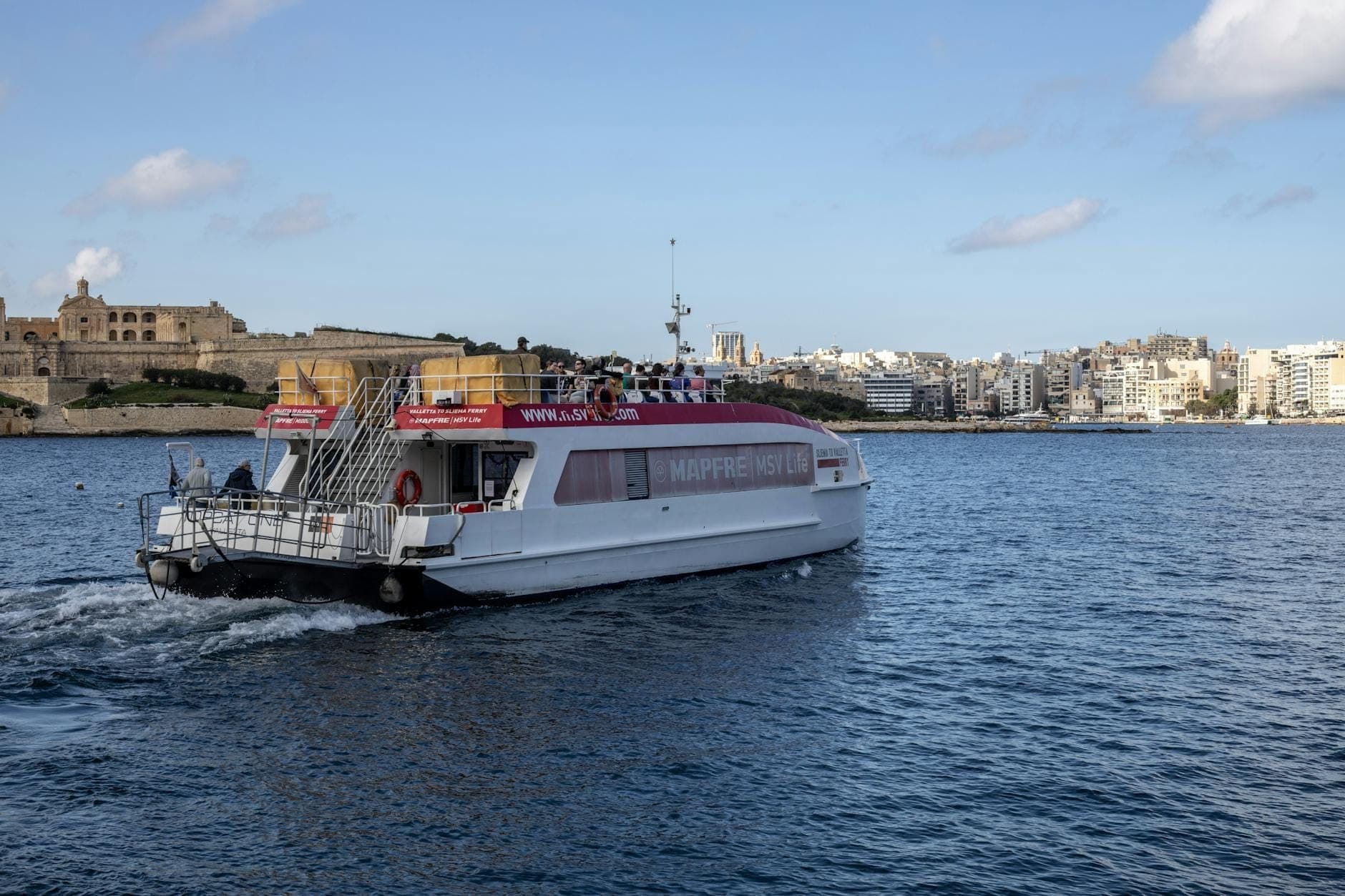 Un ferry de pasajeros cruzando un puerto en Malta con el horizonte y las fortificaciones de Valletta visibles al fondo bajo un cielo azul.