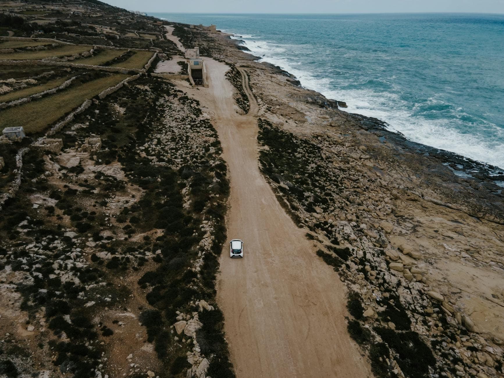 Un pequeño coche circula por una accidentada carretera costera en Malta con terreno rocoso y el mar Mediterráneo a un lado.