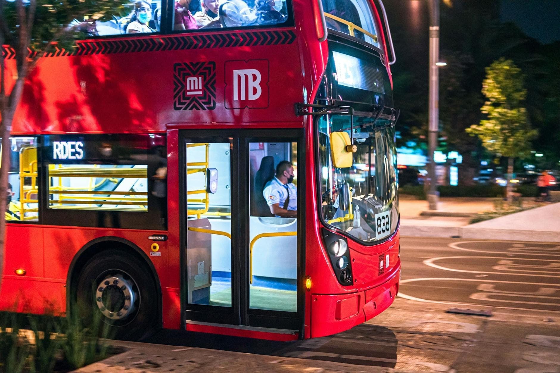 ภาพใกล้ชิดของรถบัสสองชั้นสีแดงของ Metrobús ในตอนกลางคืนที่ Mexico City โดยมองเห็นผู้โดยสารผ่านกระจกหน้าต่าง