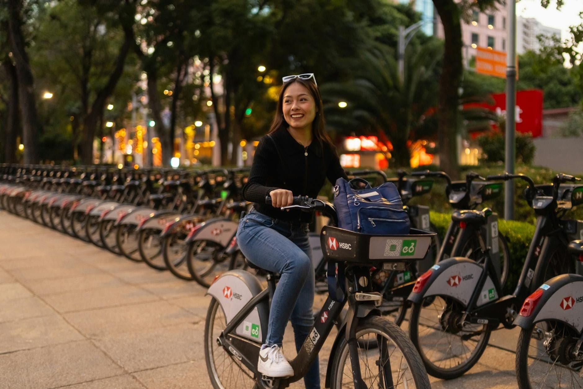 ผู้หญิงยิ้มแย้มขี่จักรยาน Ecobici ที่สถานีจอดจักรยานบนถนนร่มรื่นในเม็กซิโกซิตี้ พร้อมจักรยานจอดเรียงราย