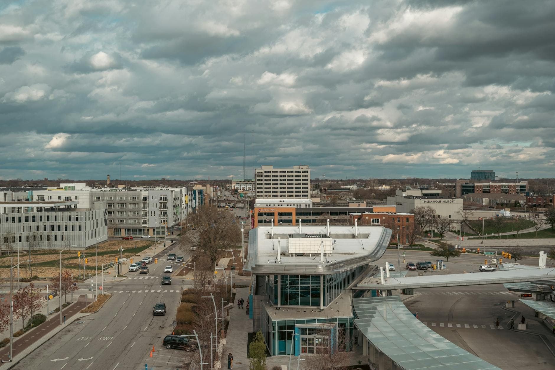 Luftaufnahme eines modernen Verkehrsknotenpunkts mit gebogenem Glasdach in einem städtischen Gebiet, umliegenden Straßen und mittelhohen Gebäuden, bewölkter Himmel darüber.