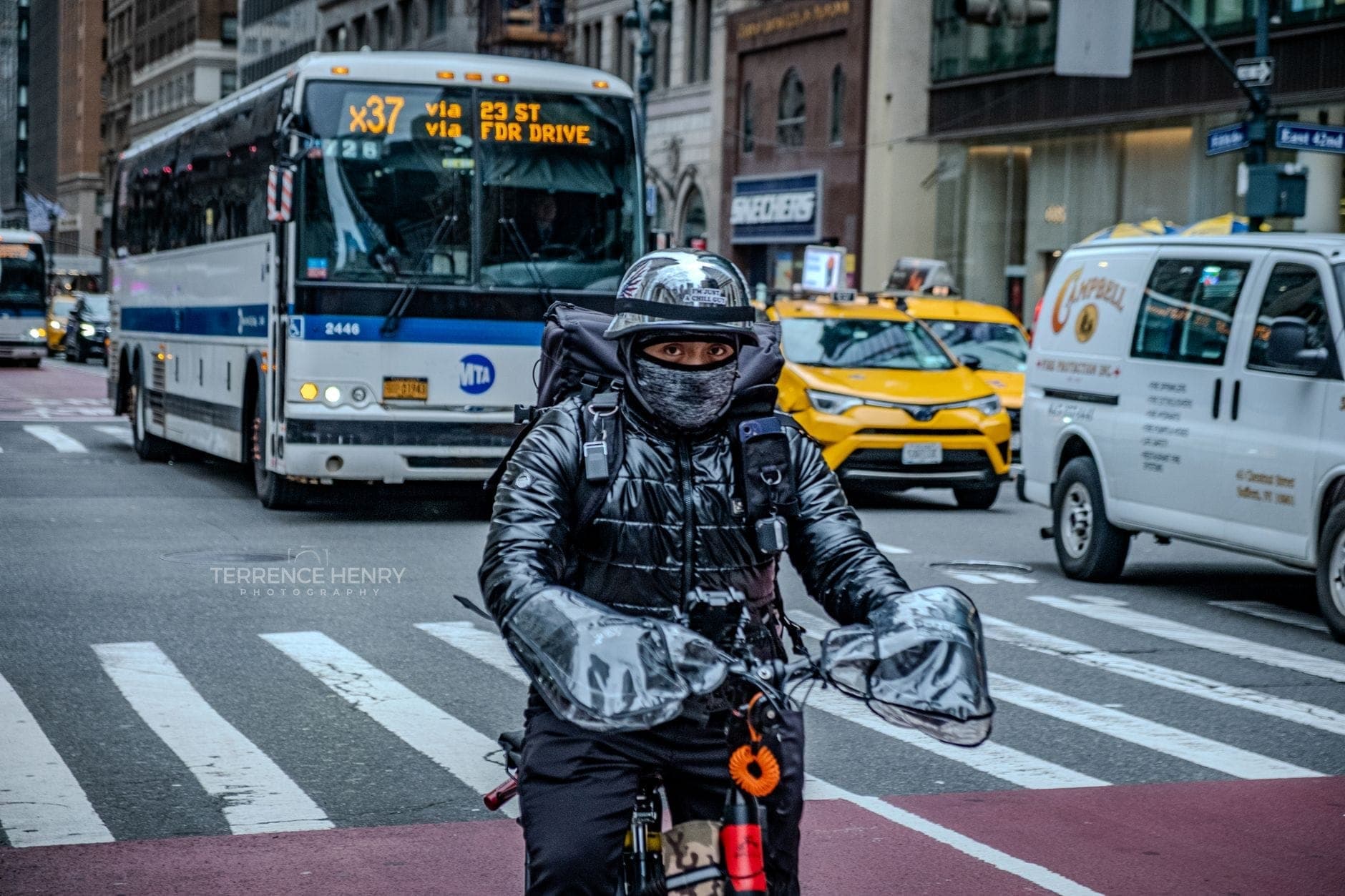 บุคคลหนึ่งขี่จักรยานบนถนนใน New York City โดยมีรถบัส MTA แท็กซี่ และอาคารในเมืองเป็นฉากหลัง