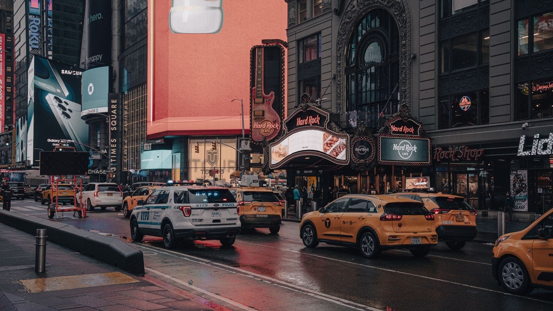 ภาพบรรยากาศที่ Times Square พร้อมแท็กซี่สีเหลืองของ NYC หลายคันขับผ่านหน้า Hard Rock Cafe และป้ายโฆษณาที่สว่างไสว