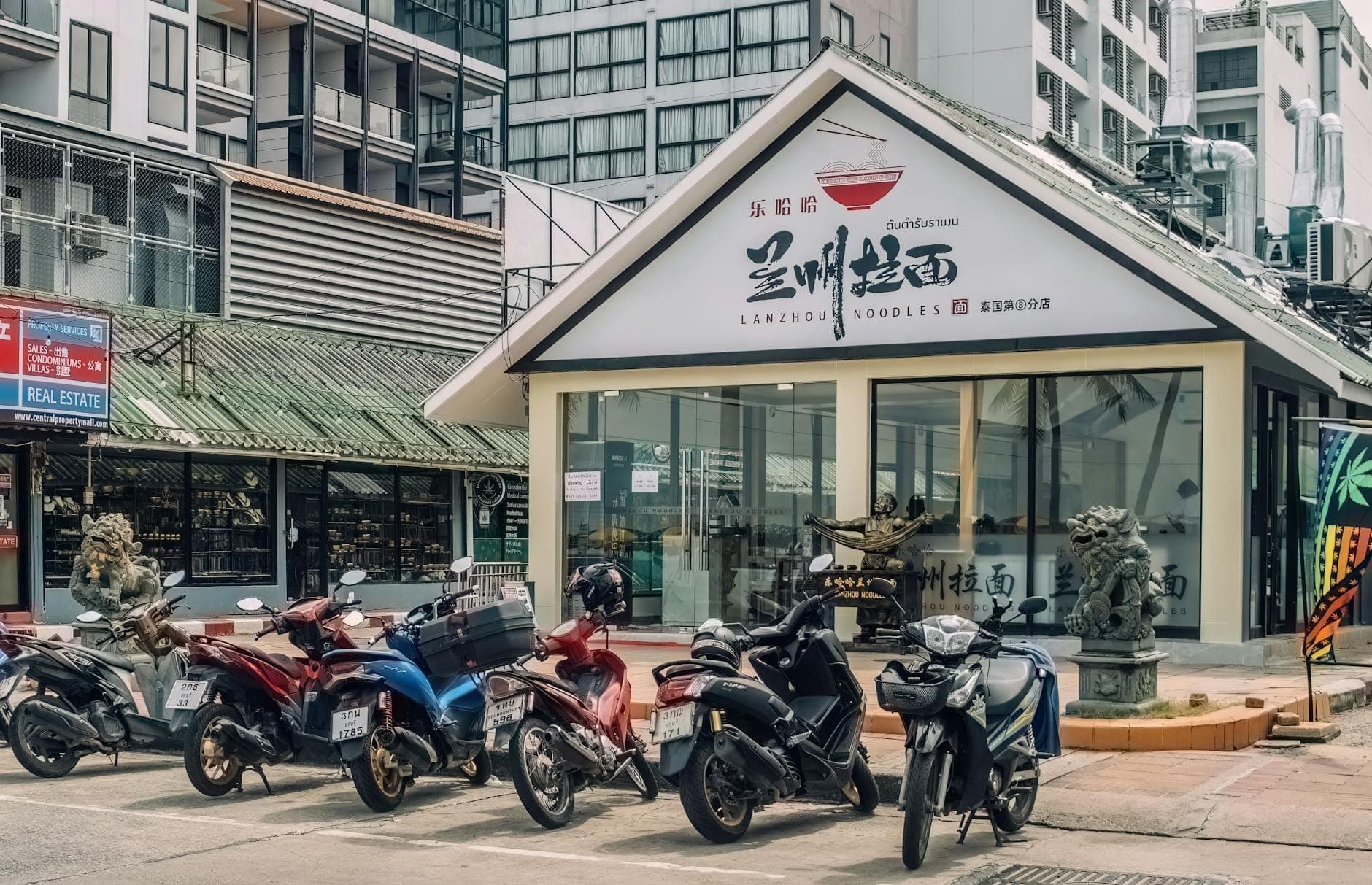 Несколько припаркованных скутеров и мотоциклов перед витриной магазина на современной городской улице в Паттайе.