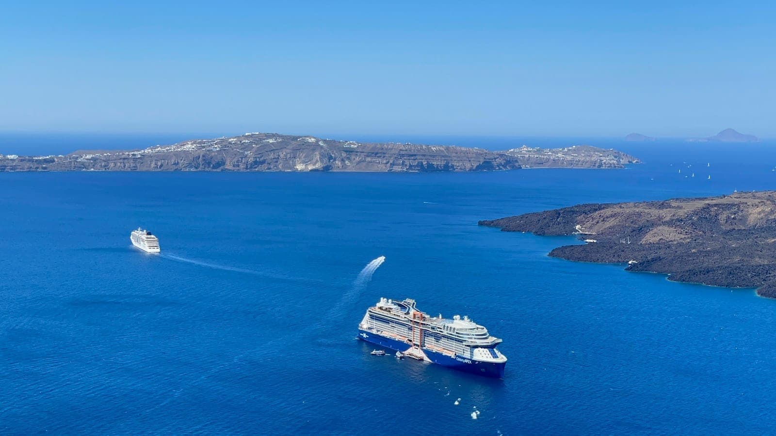 Luftaufnahme der Caldera von Santorini mit mehreren Fähren und Kreuzfahrtschiffen, die über blaues Wasser und Vulkaninseln anlegen.