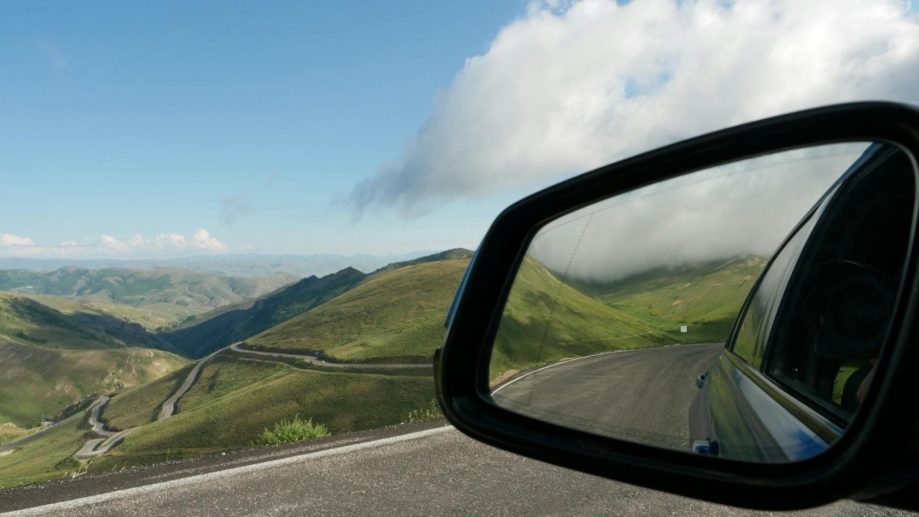 Pemandangan dari kaca spion samping mobil yang memperlihatkan jalan pegunungan berkelok-kelok dan bukit hijau di bawah langit berawan sebagian di Sisilia.