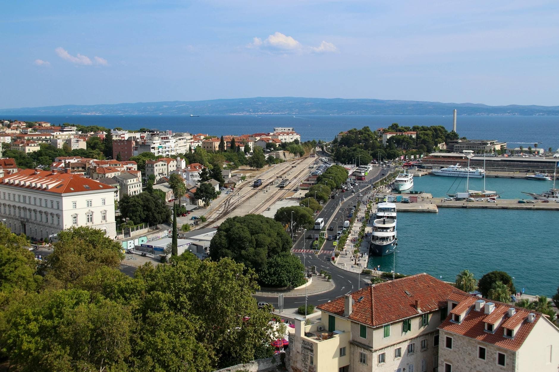 Vista aerea di Spalato con i binari ferroviari, la stazione degli autobus e il porto dei traghetti sul mare Adriatico, evidenziando le principali opzioni di trasporto.