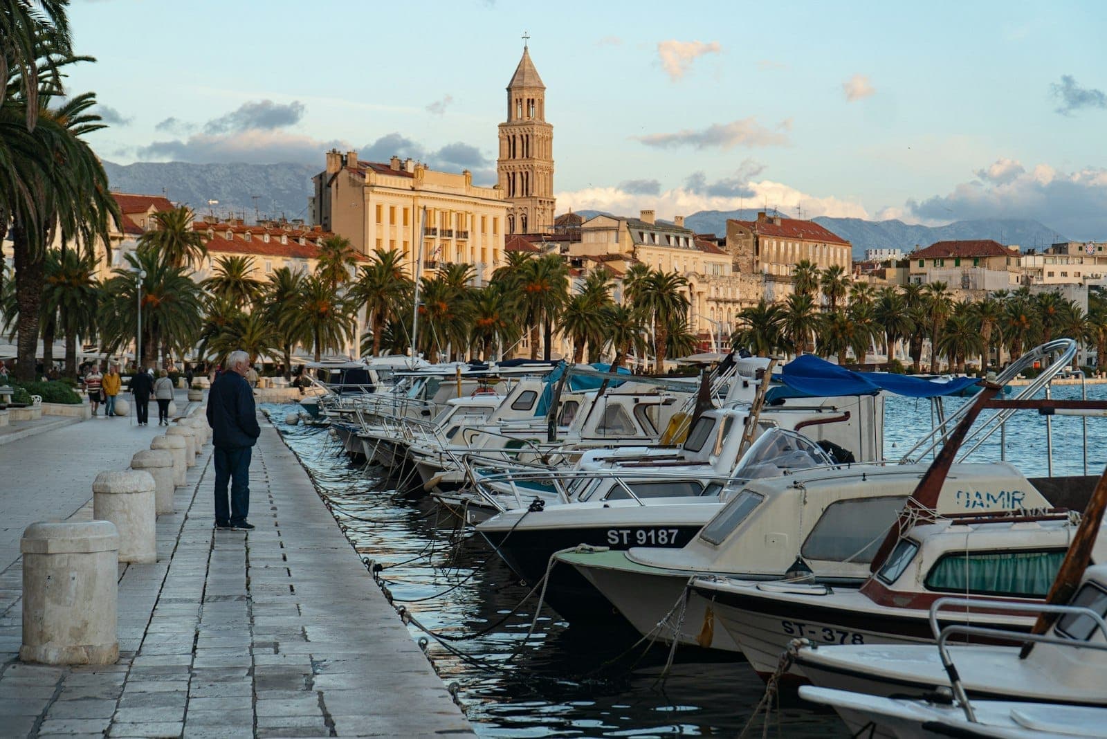 Barche ormeggiate lungo la passeggiata Riva di Split, con palme e il campanile del Palazzo di Diocleziano sullo sfondo al tramonto.