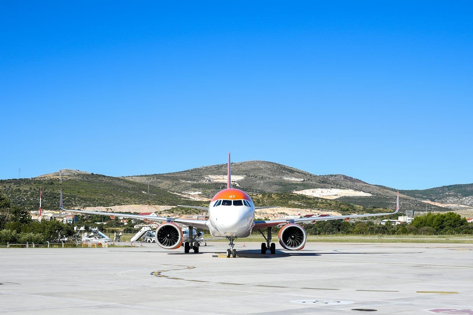 Un aereo commerciale sul piazzale di un aeroporto con colline sullo sfondo sotto un cielo azzurro limpido.