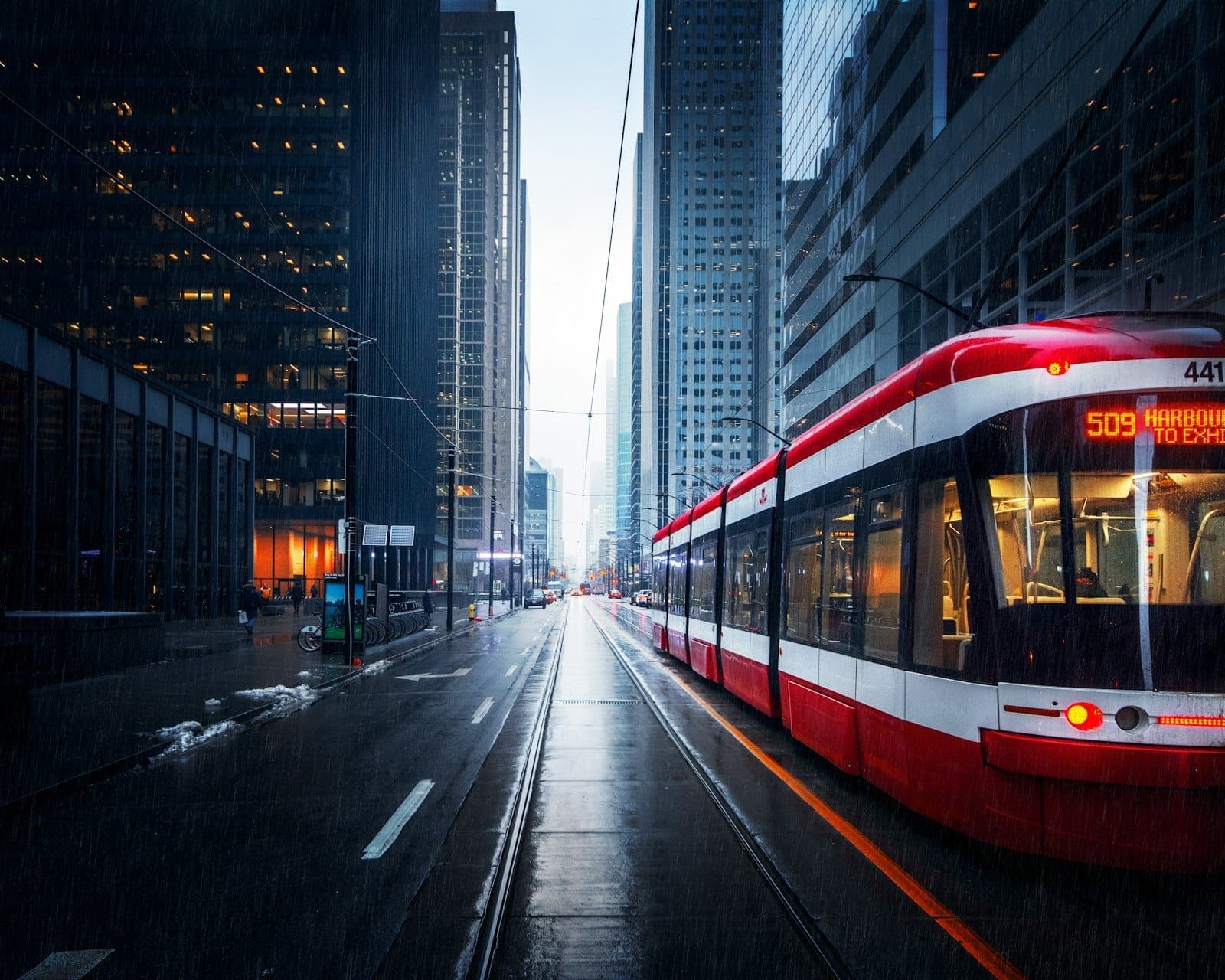 Sebuah streetcar Toronto berwarna merah dan putih melaju di jalan pusat kota yang diapit gedung-gedung kaca bertingkat tinggi di hari hujan.